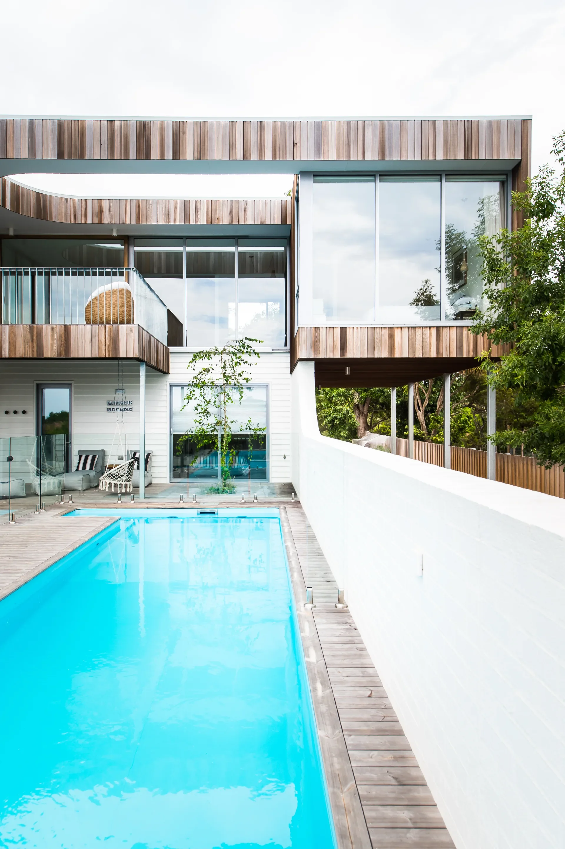 This modern home features a long lap pool set into a wooden deck and bordered by a white brick wall. The building above uses vertical wooden cladding and large glass windows, with a covered balcony overlooking the water.