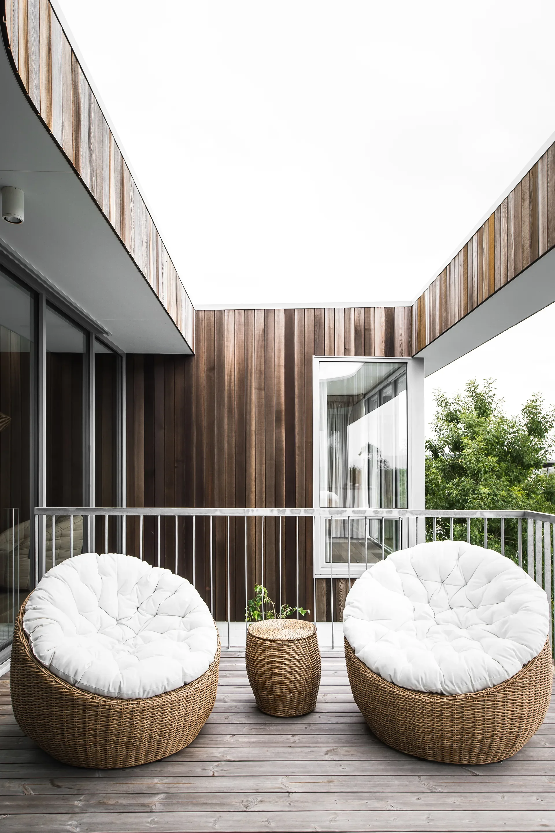 Two round wicker chairs with white cushions sit on a wooden balcony under a large square cutout in the ceiling. The balcony is surrounded by a metal railing and vertical wood paneling.