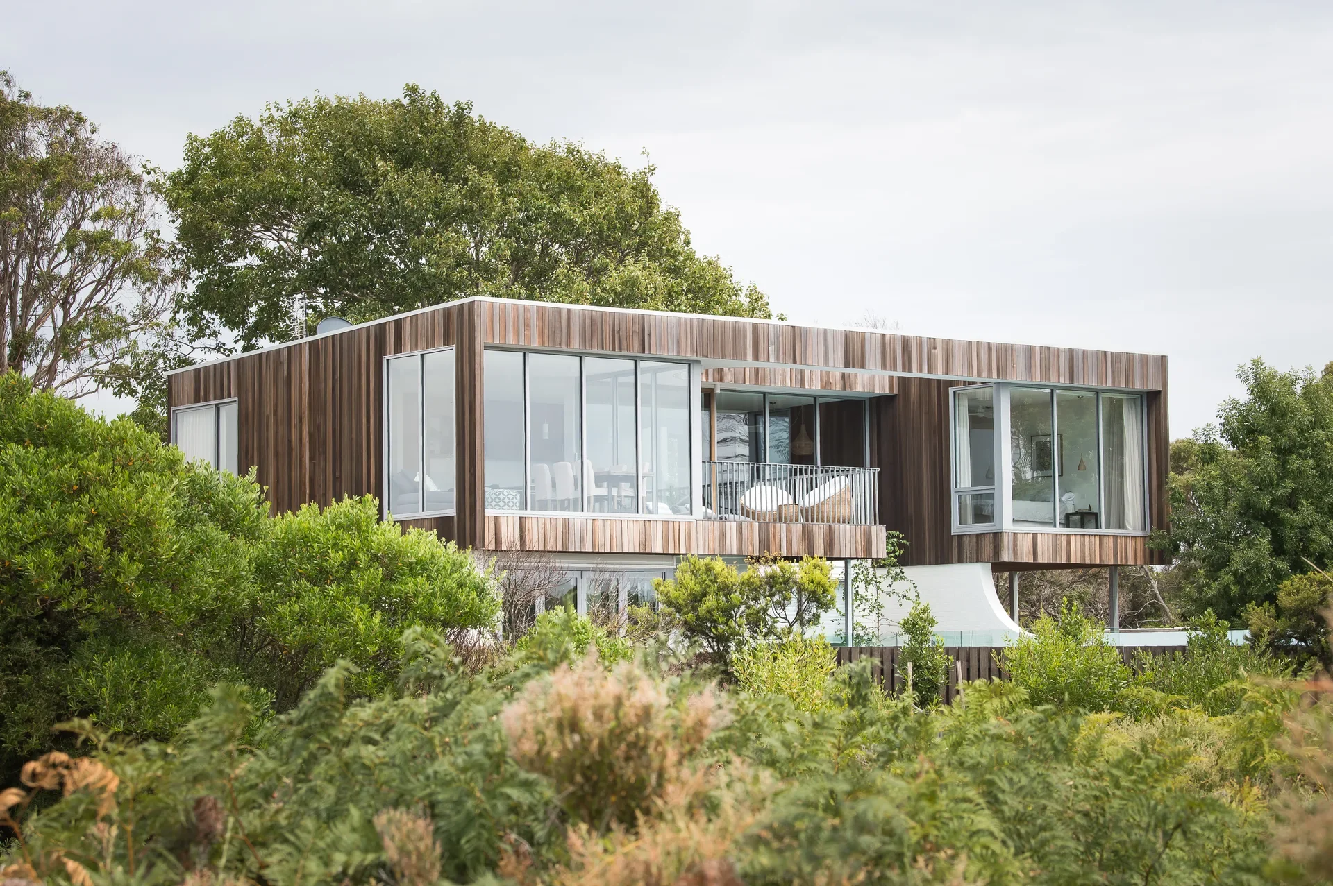 This modern, rectangular home is clad in vertical wood siding and features large floor-to-ceiling glass windows. It sits partially hidden by dense green shrubs and trees.