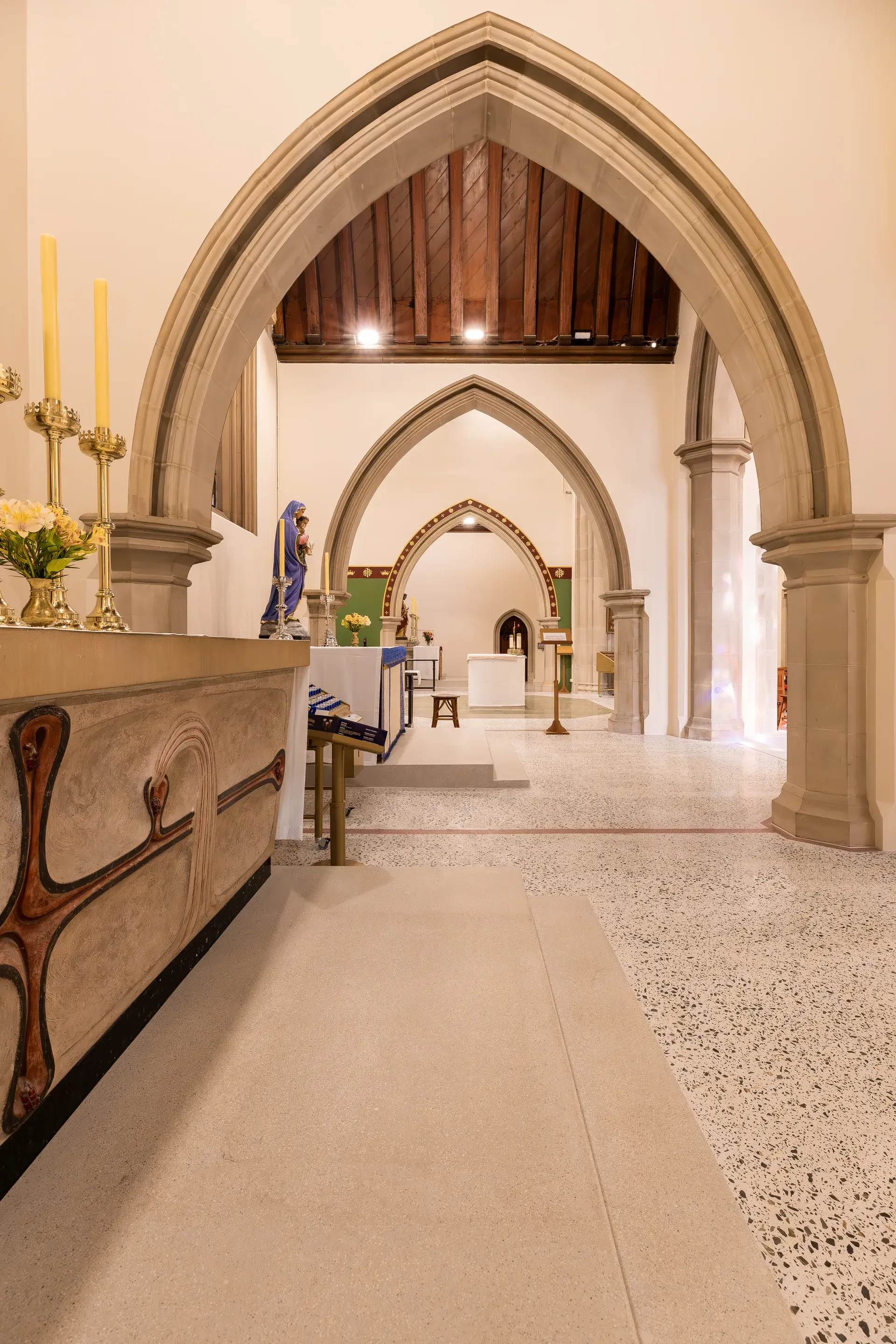Interior view of the cathedral featuring a series of stone pointed arches leading toward an altar, with a timber-beamed ceiling and speckled stone flooring.