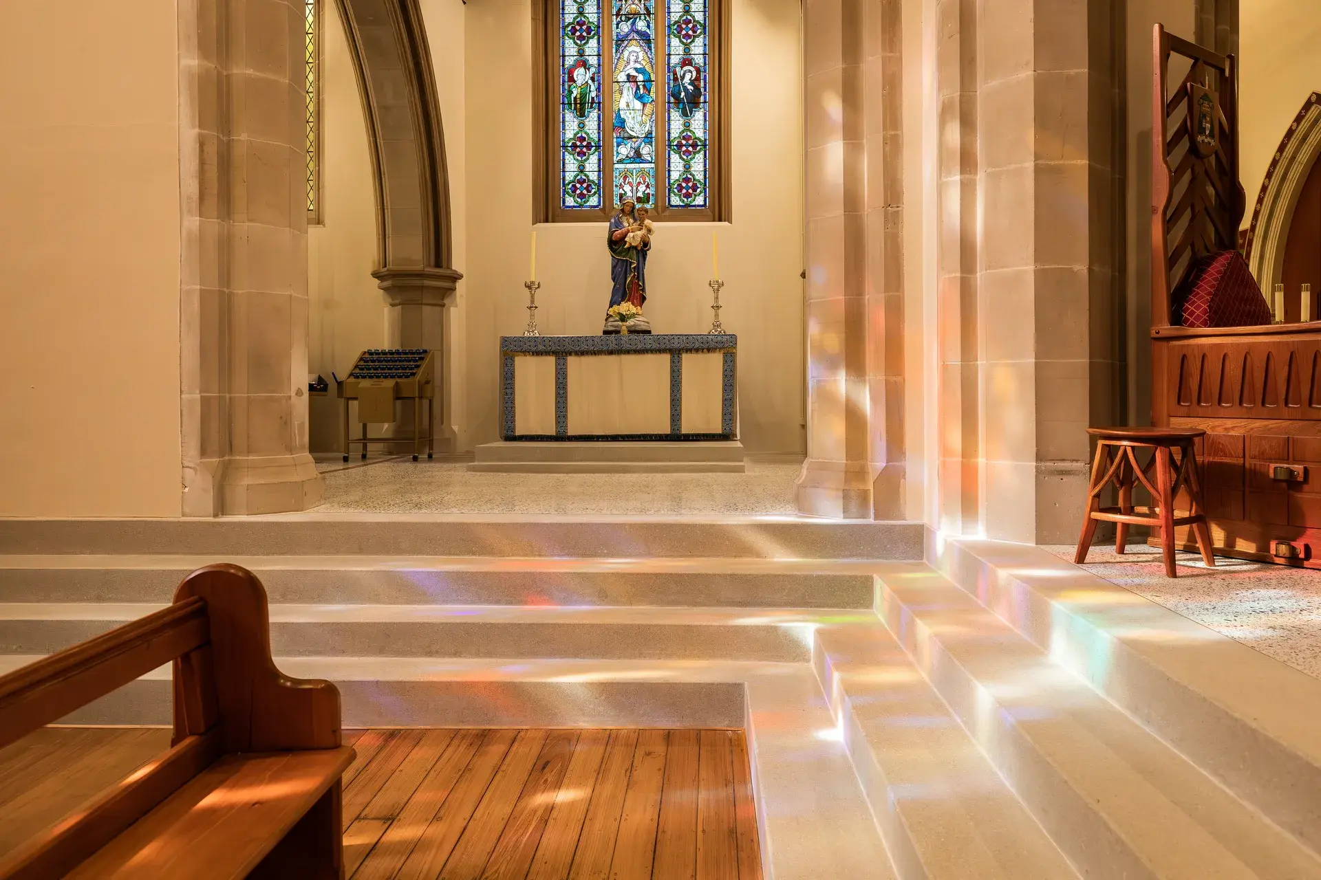 Interior view of a church side chapel featuring stone steps illuminated by rainbow light from a stained-glass window, leading to an altar with a statue depicting religious figures.
