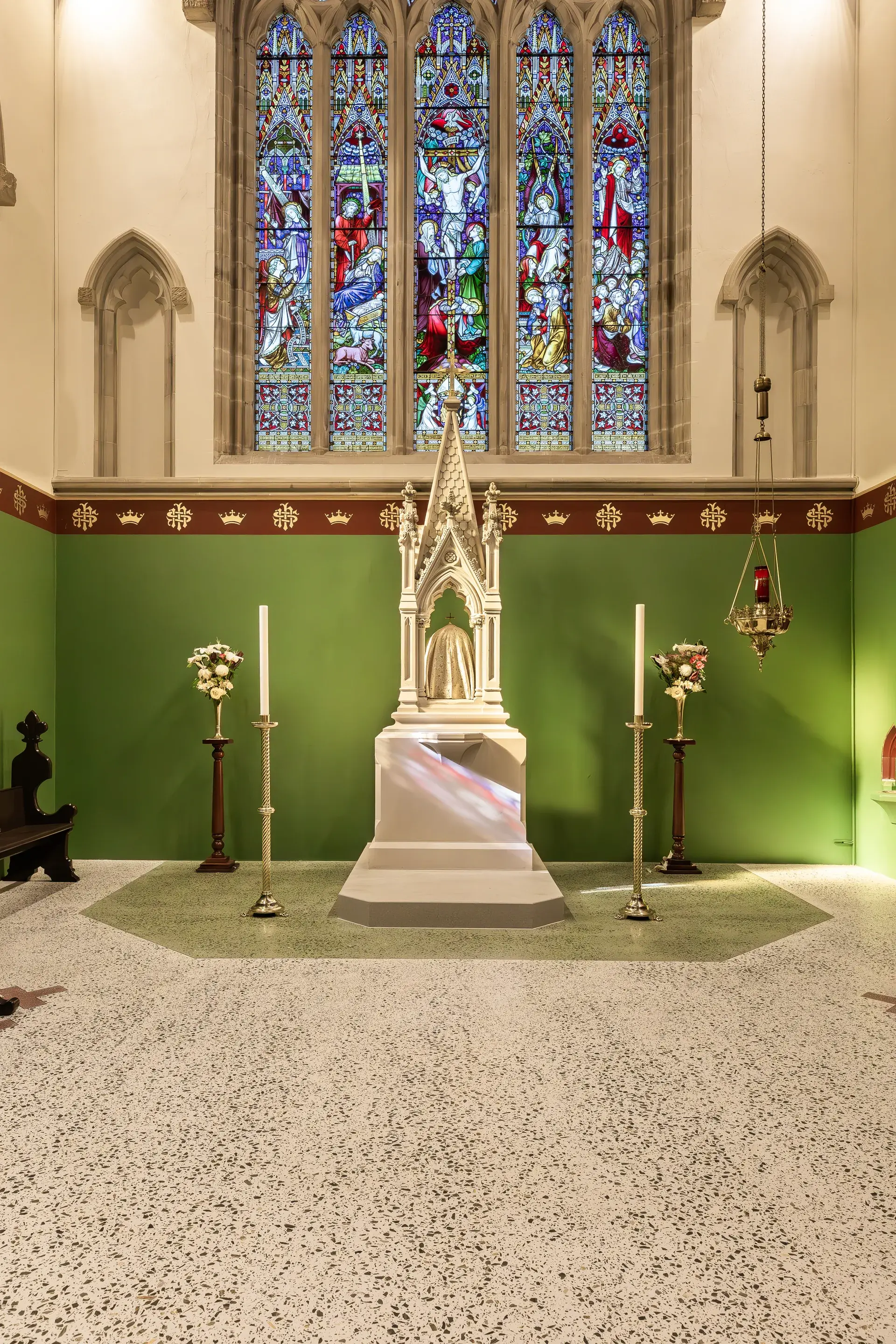 An altar featuring an ornate white tabernacle centered against a green wall, positioned beneath a large stained-glass window depicting religious figures.