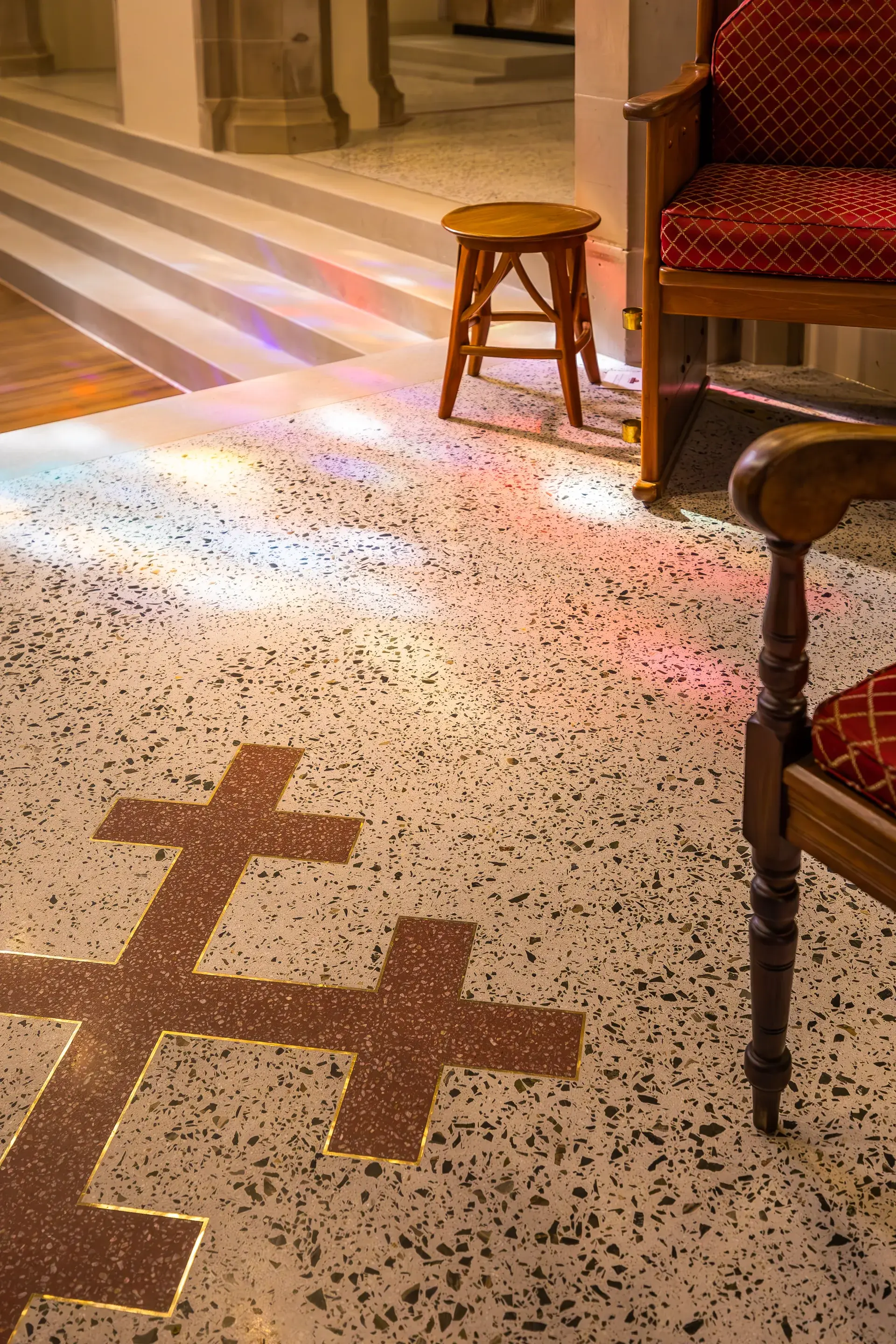 A close-up of a speckled stone floor featuring a large inlaid cross, with colourful light from stained-glass windows reflecting across the surface near wooden chairs and stone steps.