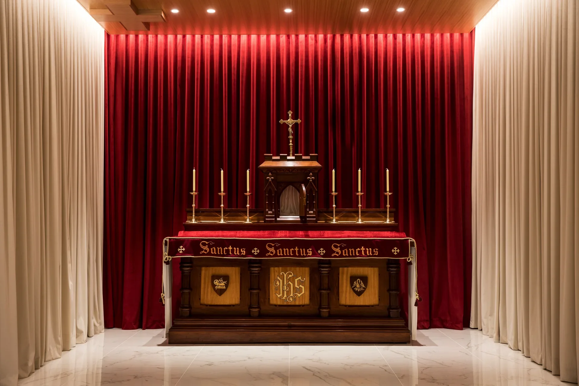 An ornate wooden altar stands against a vibrant red curtain in a brightly lit sanctuary. It features several tall candles, a central cross, and the repeated inscription "Sanctus" across its velvet-covered front.