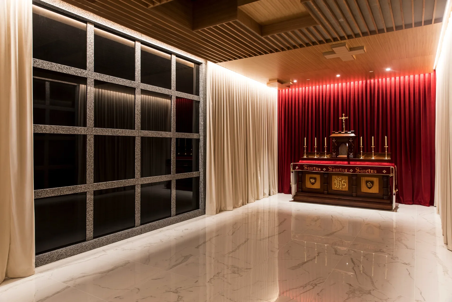 This sanctuary features a marble floor and a wooden slat ceiling with recessed cross motifs. An ornate altar stands before a vibrant red curtain, flanked by cream drapes and a dark, grid-patterned glass wall.