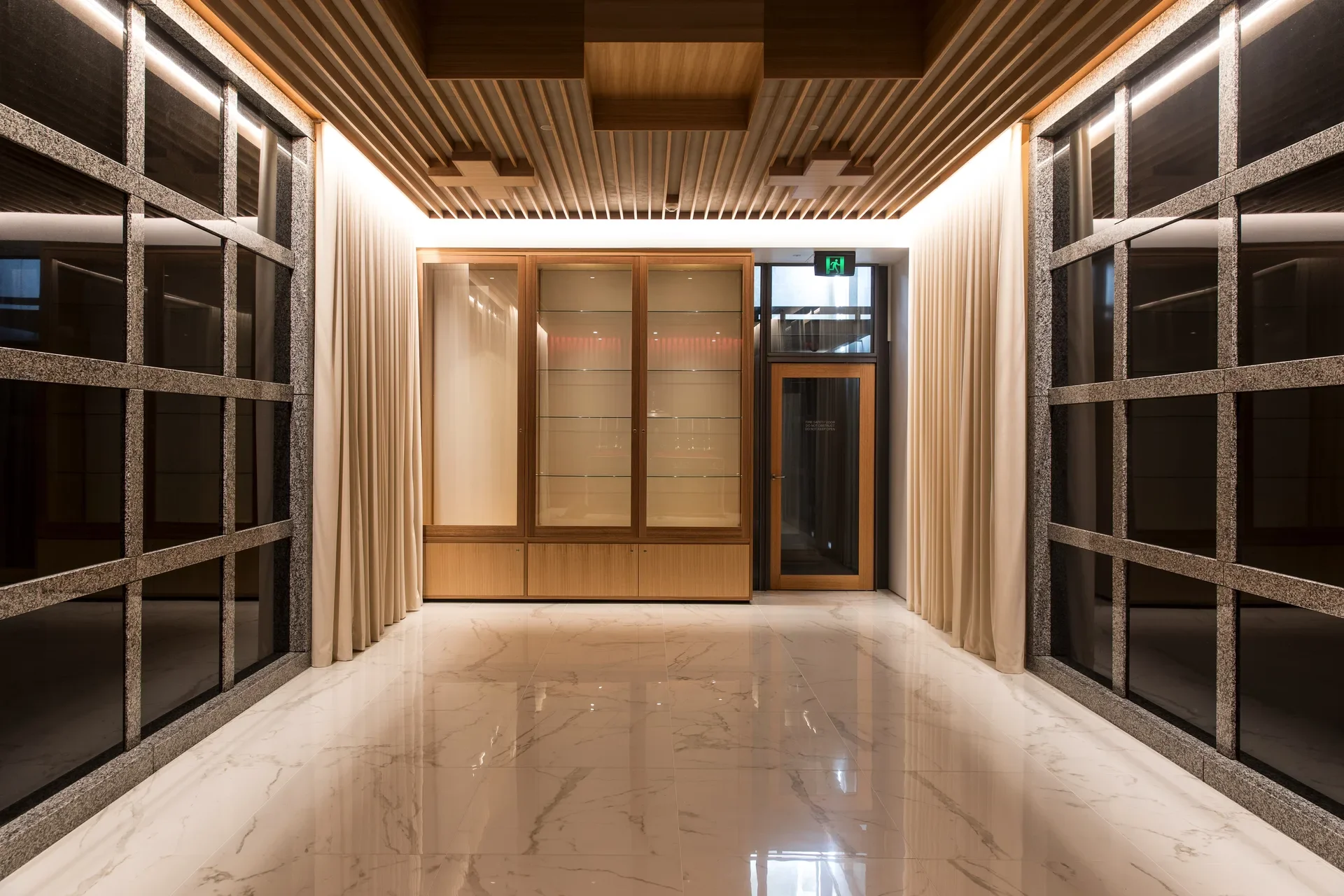 A hallway features a glossy marble floor and a wooden slat ceiling. It is lined with cream drapes and dark, grid-patterned glass panels.