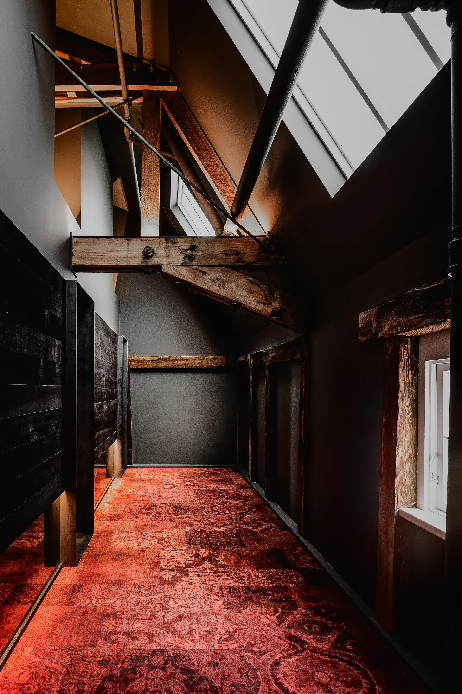 This narrow hallway features heavy timber beams and dark grey walls beneath a slanted ceiling with a skylight. The floor is covered in a vibrant red patterned carpet, and the walls are accented with dark wood paneling and black metal pipes.