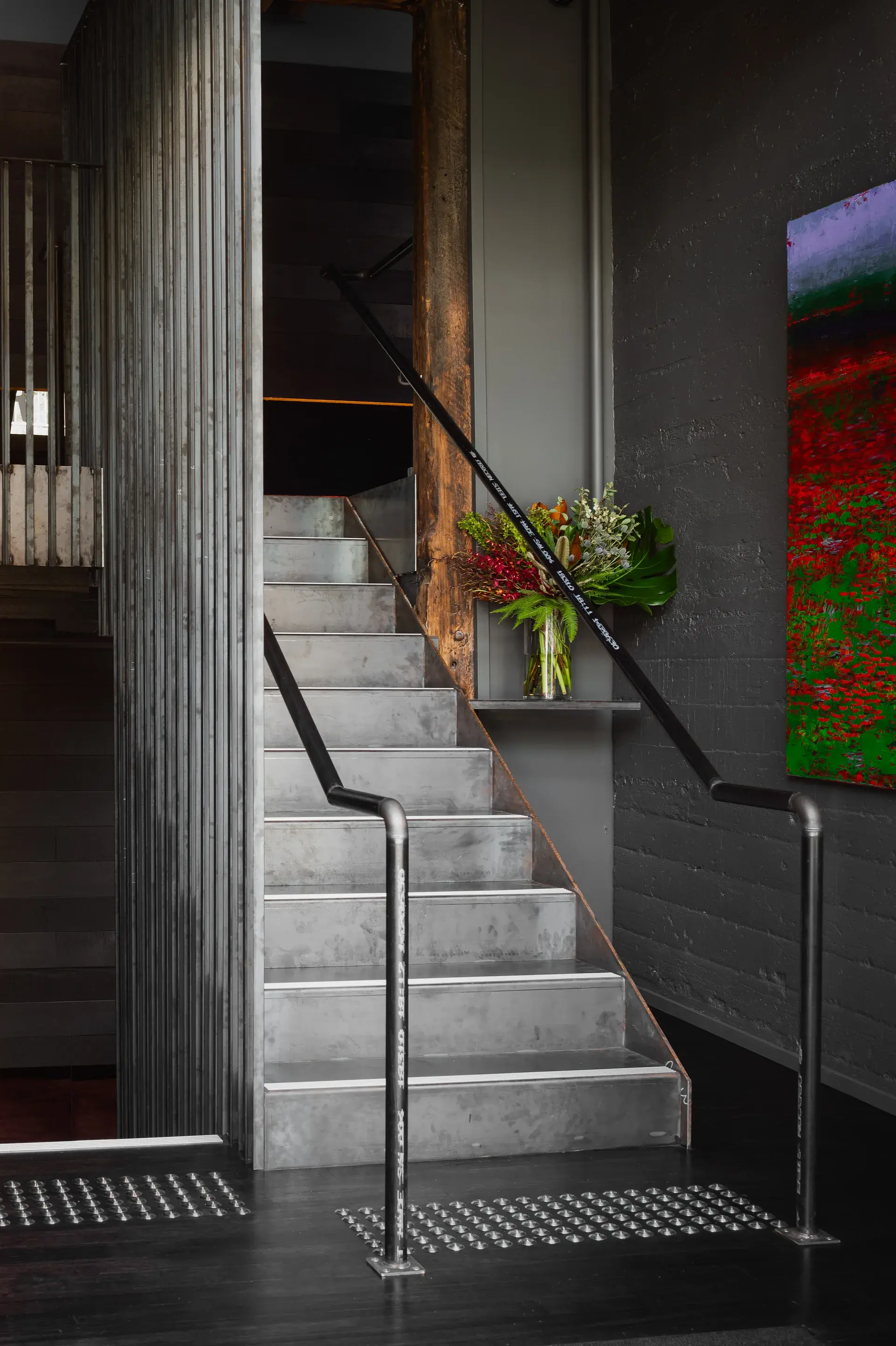 This industrial-style staircase is made of galvanised metal and features a sleek black handrail. It is set against a dark grey textured wall adorned with a vibrant, colourful painting and a small shelf holding a floral arrangement.