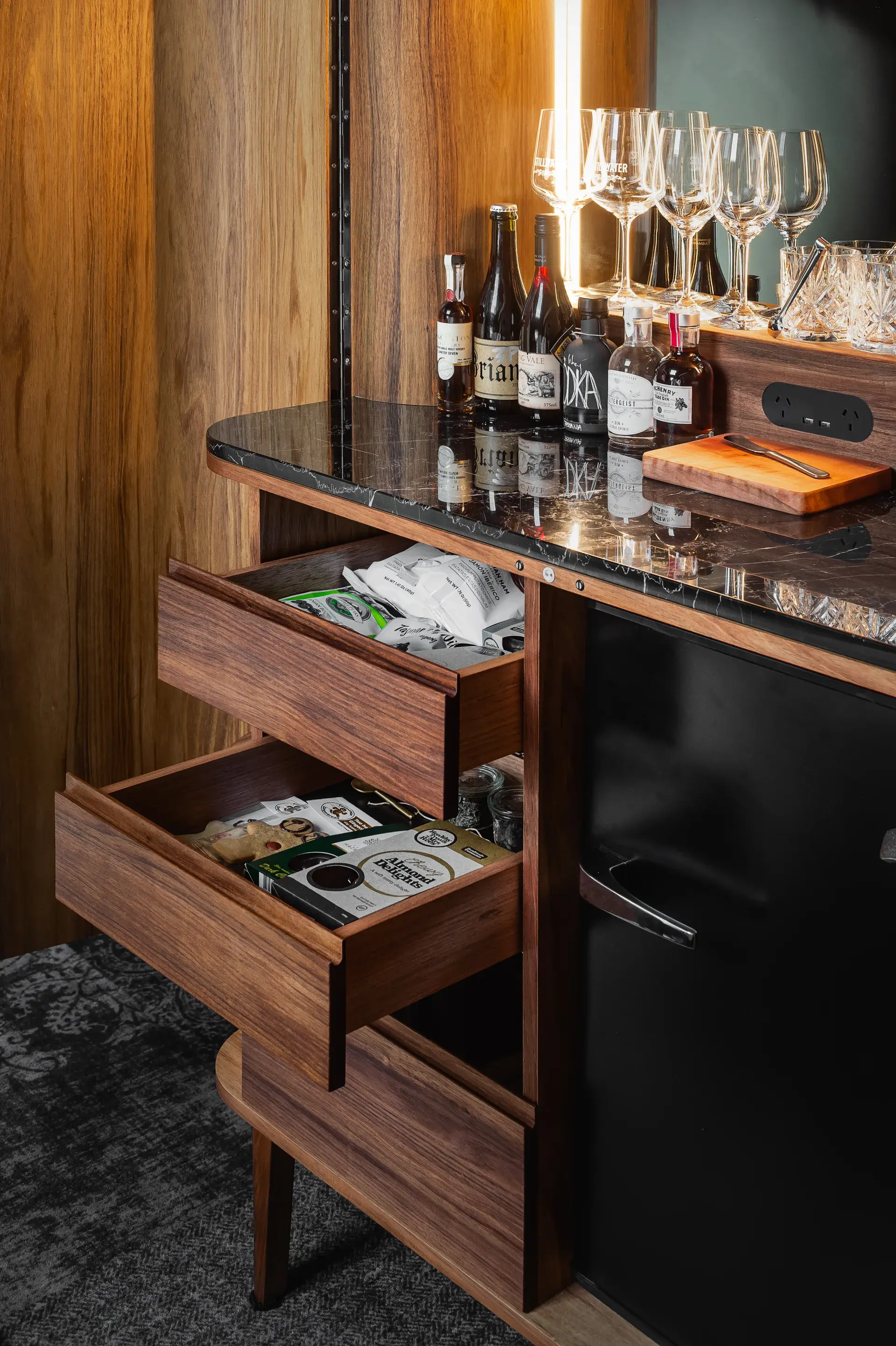 A wooden bar cabinet features a black marble top and a mirrored backsplash reflecting glassware. It includes a small black refrigerator and open drawers filled with snacks and drinks.