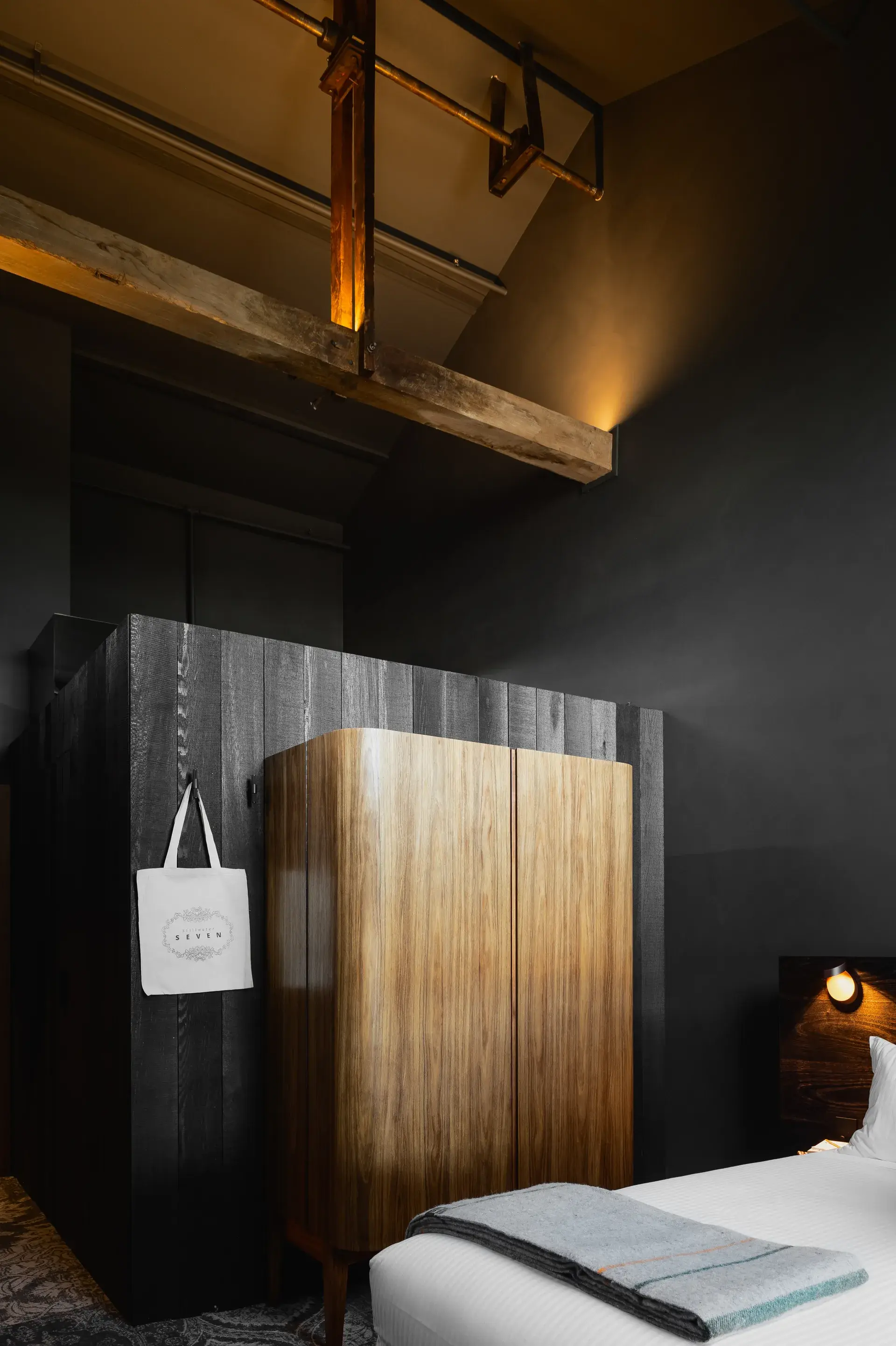This moody bedroom features a curved wooden wardrobe set against a black panelled wall. The space is characterised by exposed timber beams and dark grey walls, with a white bed and a small glowing sconce in the corner.