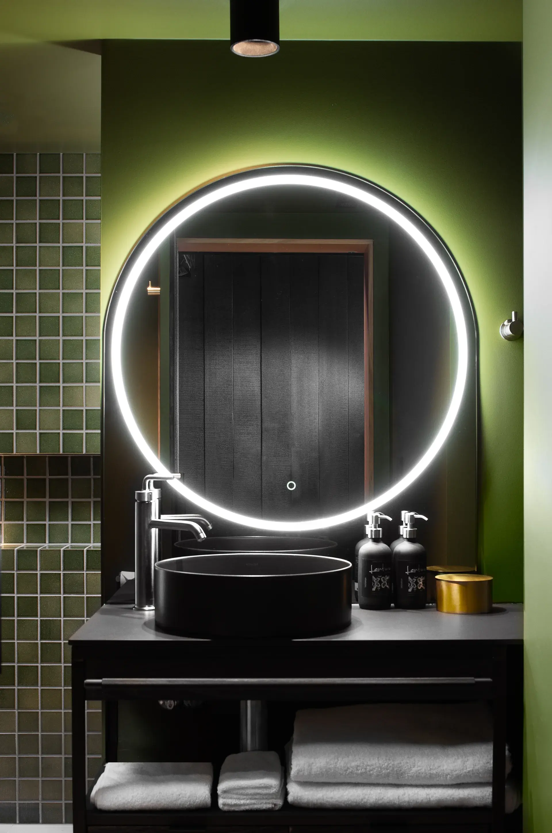 This bathroom features olive-green walls and a vanity with a black vessel sink and a large, circular backlit mirror. To the left, a wall of small green tiles adds texture to the space, while black accessories and white towels complete the modern look.
