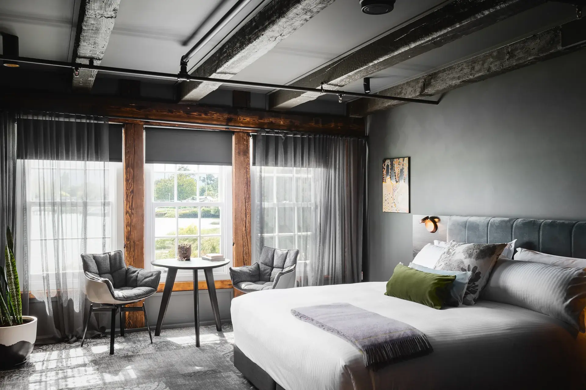 This industrial-style bedroom features exposed timber beams across a grey ceiling and dark grey walls. A small seating area with two grey chairs and a round table is positioned by windows framed in natural wood, while the bed is accented with a grey velvet headboard.