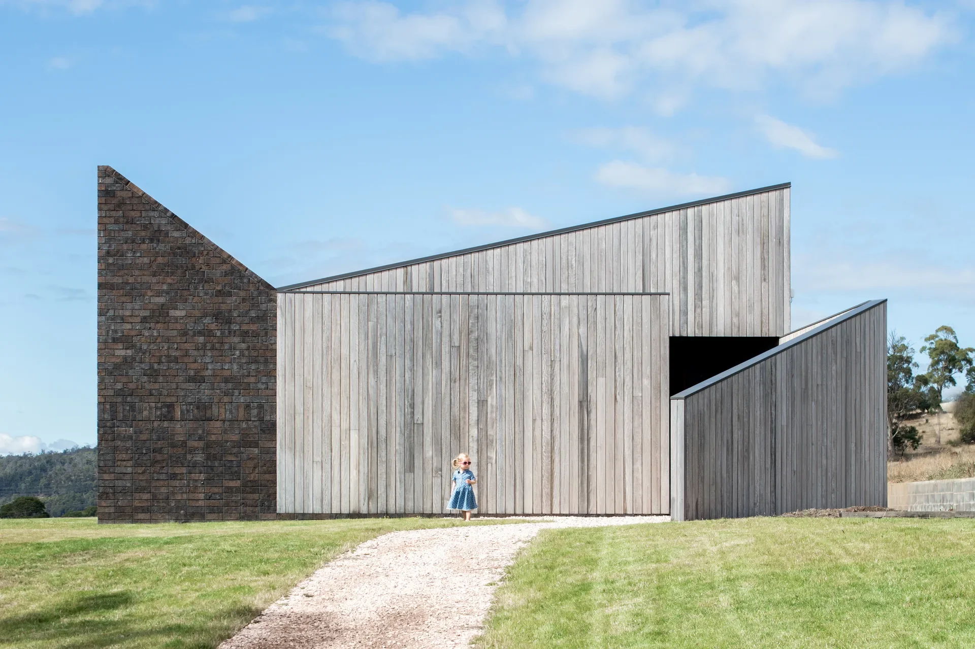 This modern cellar door features sharp, geometric lines with a mix of grey vertical wood siding and a dark, textured brick section. A gravel path leads toward the building across a grassy field under a bright blue sky.