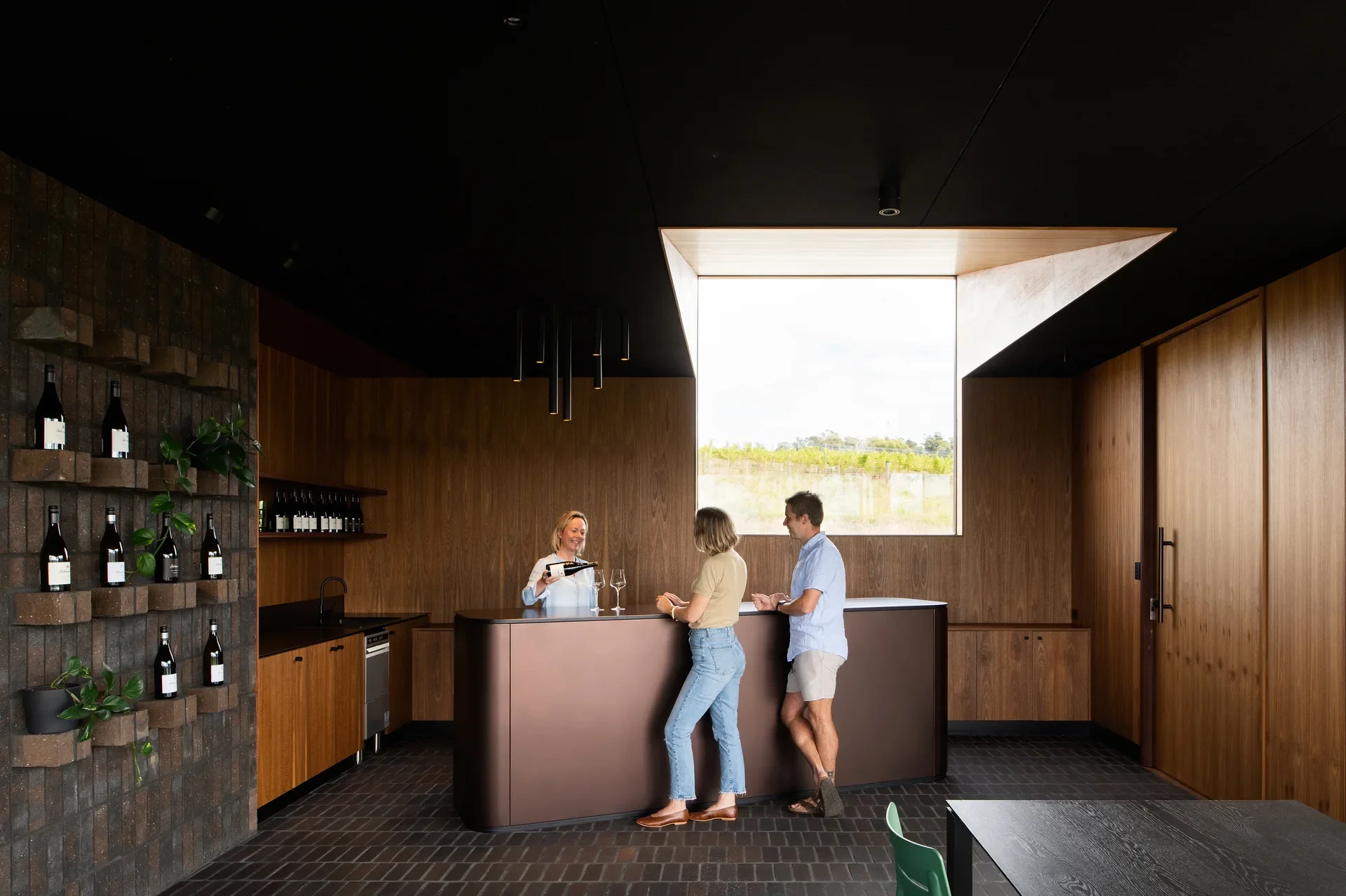 A sleek copper-toned bar sits in a tasting room with warm wood paneling and dark brick floors. A server pours wine for a couple near a large window overlooking a vineyard.