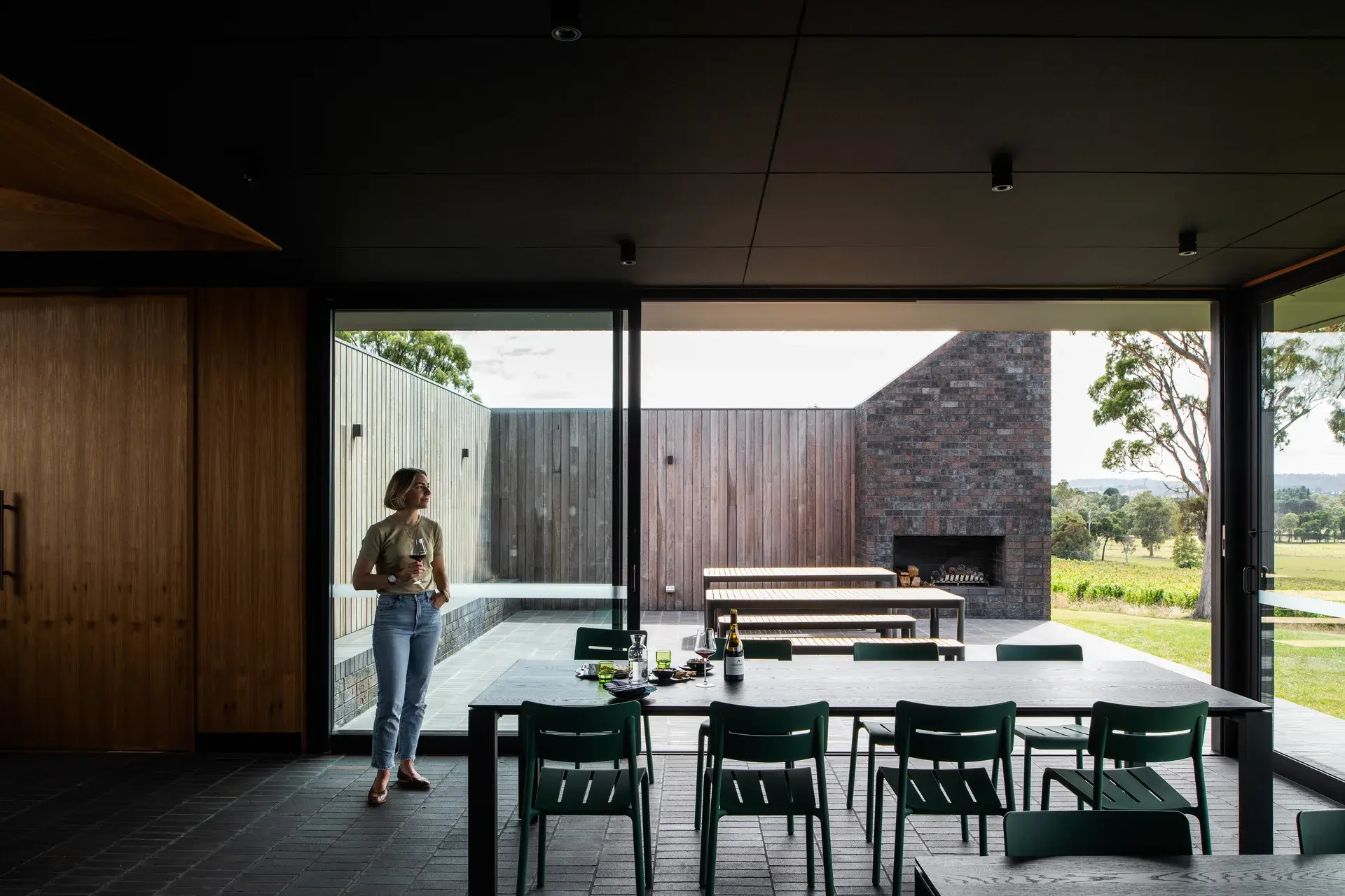 A woman stands in a dining area holding a glass of wine, looking out through large sliding glass doors. The interior features a black ceiling and dark brick floors, while the outdoor patio includes wooden picnic tables and an integrated brick fireplace.