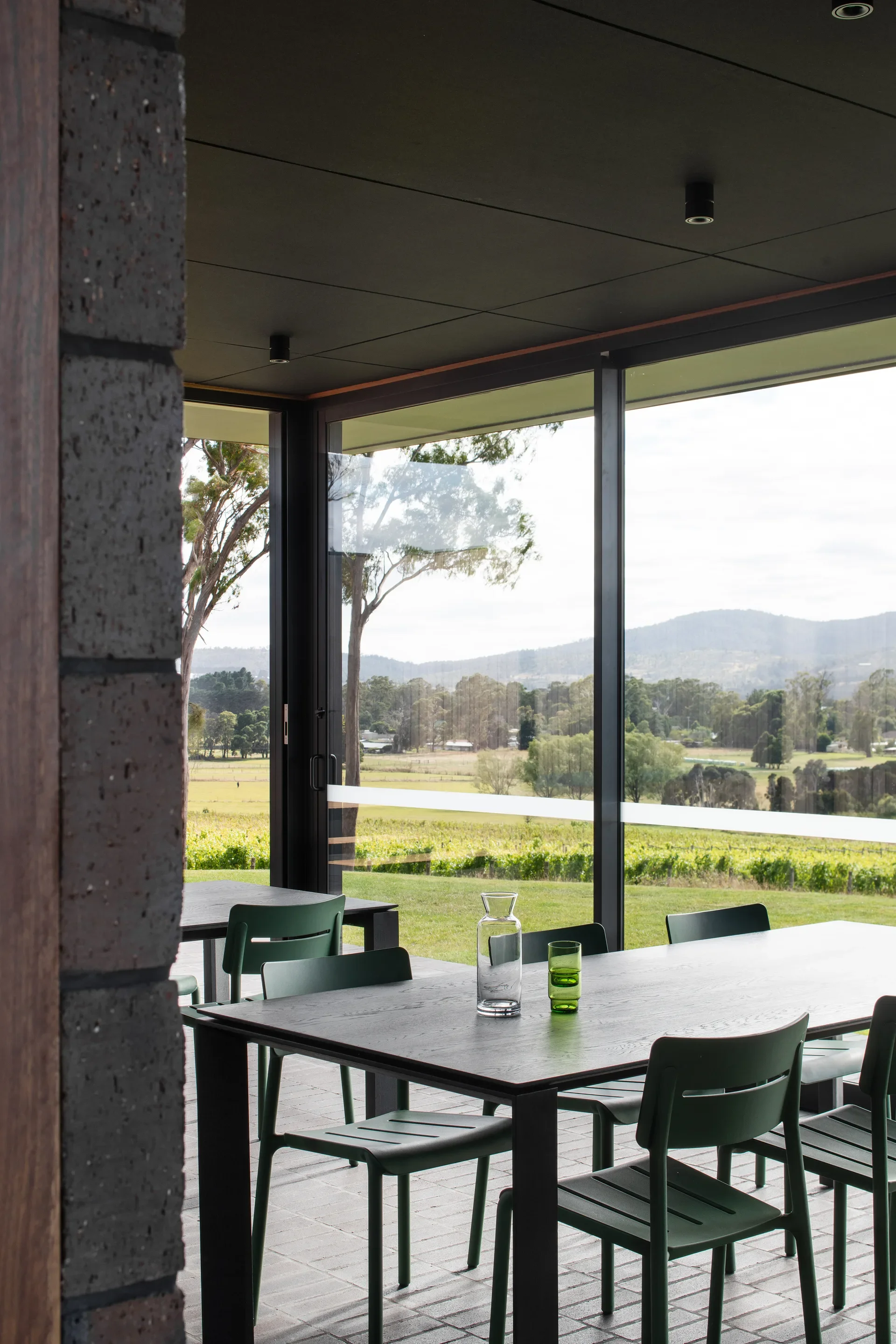 A black dining table with green chairs sits before large windows overlooking a lush vineyard and distant hills. The space features a dark ceiling with recessed spotlighting and dark brick flooring that extends toward the exterior.