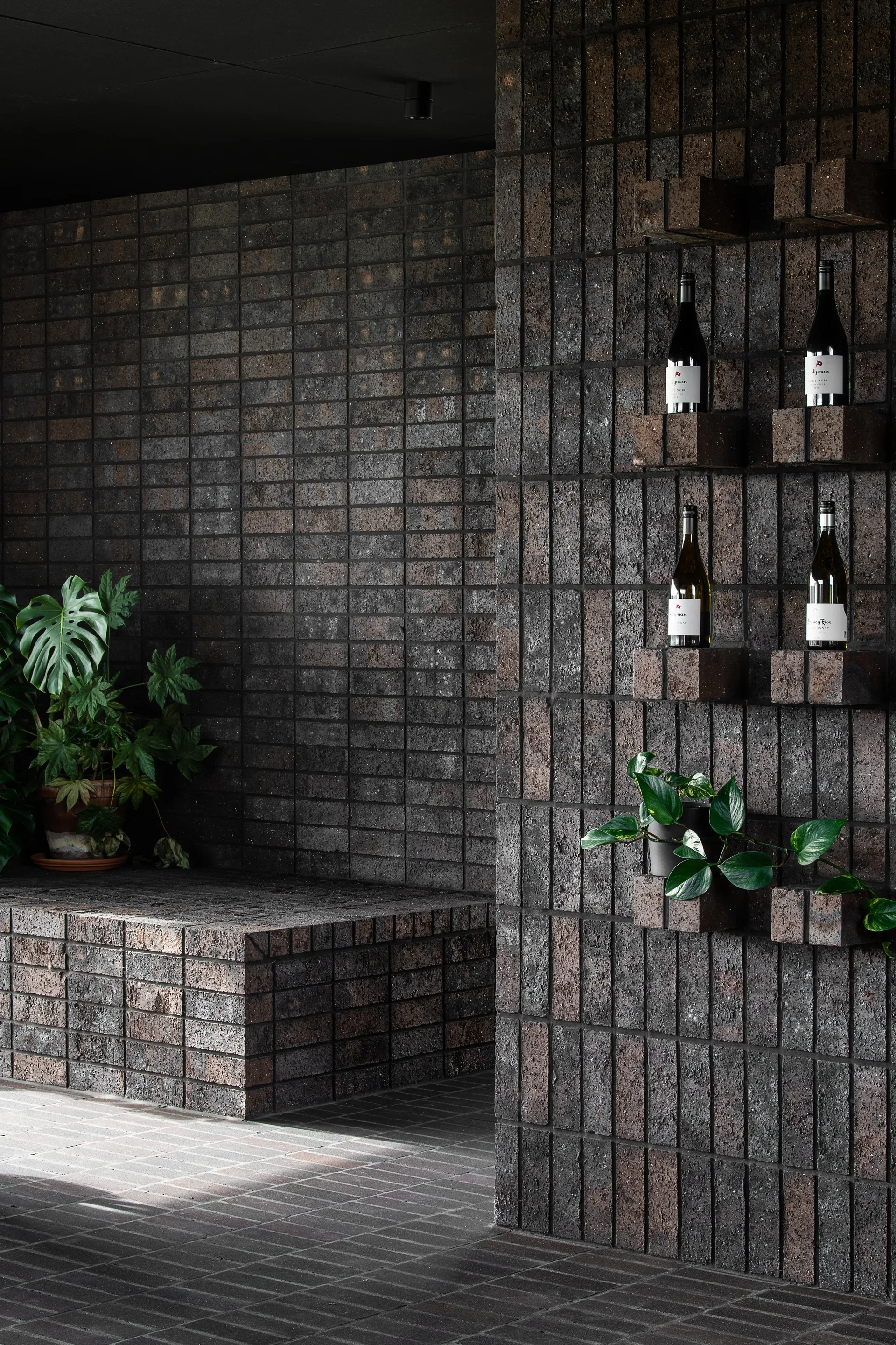 A dark, textured brick wall features wine bottles and small plants resting on individual protruding bricks. The space is finished with dark brick flooring and includes a low brick ledge accented by a large potted Monstera plant in the background.