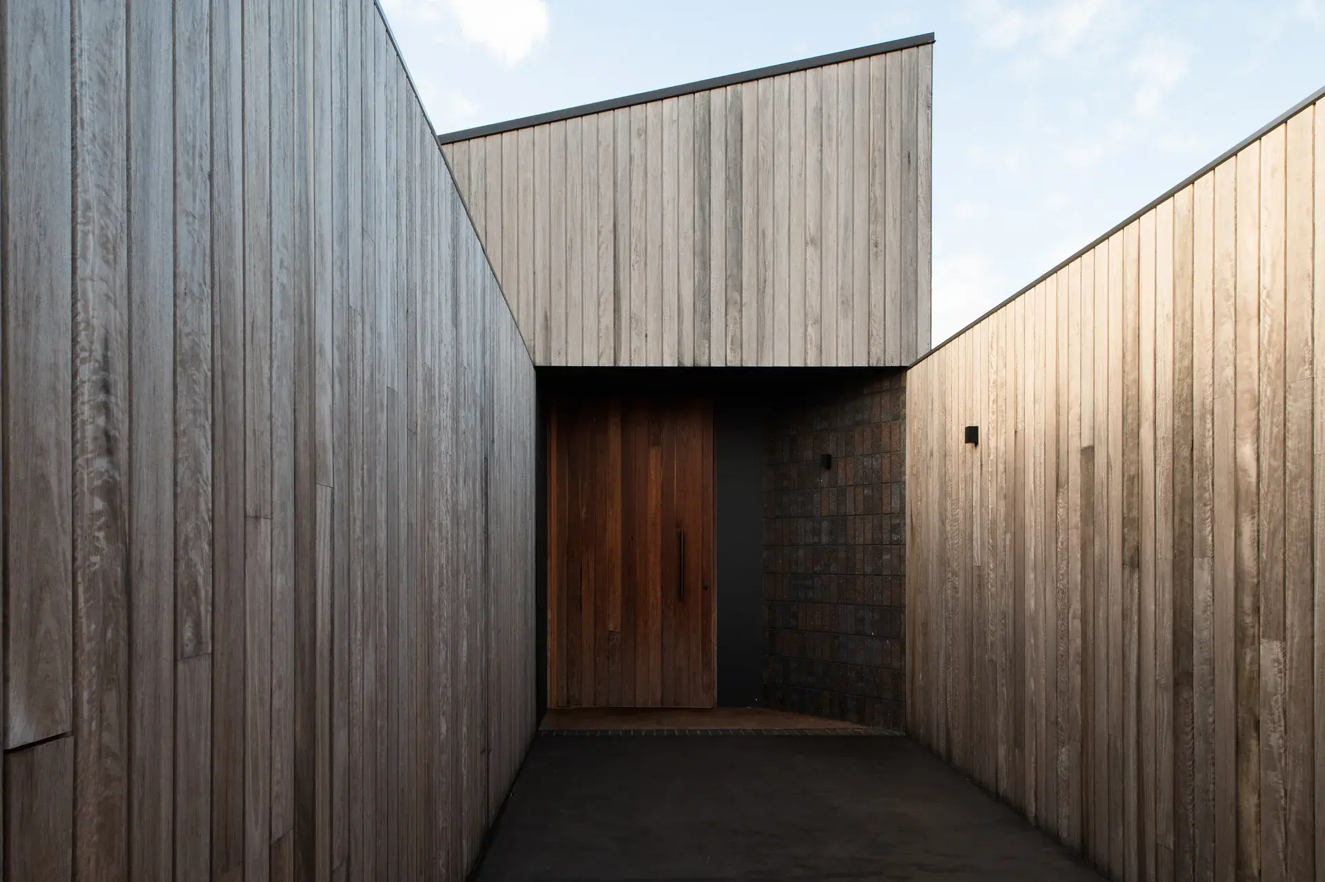 This modern entrance is recessed between high walls of vertical grey wood siding. It features a large, solid natural wood door set against a dark textured brick section.