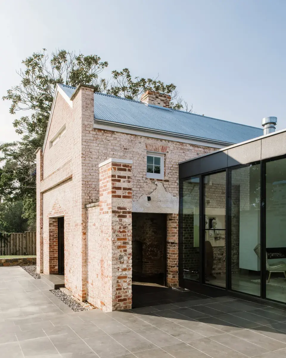 This historic brick building features a gabled roof and a prominent brick chimney structure. It is seamlessly connected to a modern, glass-walled extension with a dark frame, overlooking a grey stone patio.