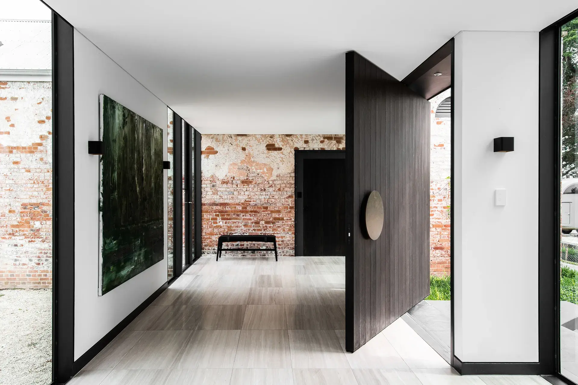 The hallway features a large, dark wood pivot door with a distinctive circular handle. The interior design pairs white walls and light tiled floors with a rugged, exposed brick wall and a large dark green abstract painting.