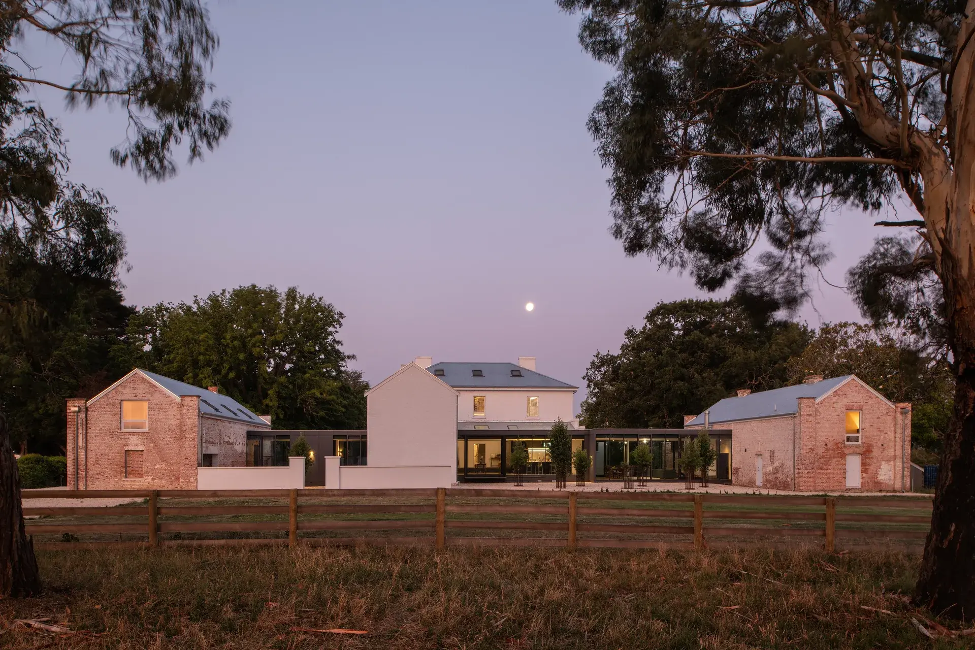 This sprawling property features a cluster of white-washed and weathered brick buildings unified by a sleek, modern glass pavilion. Seen at dusk under a full moon, the structures are surrounded by a wooden fence and large trees, with warm light glowing from the windows.