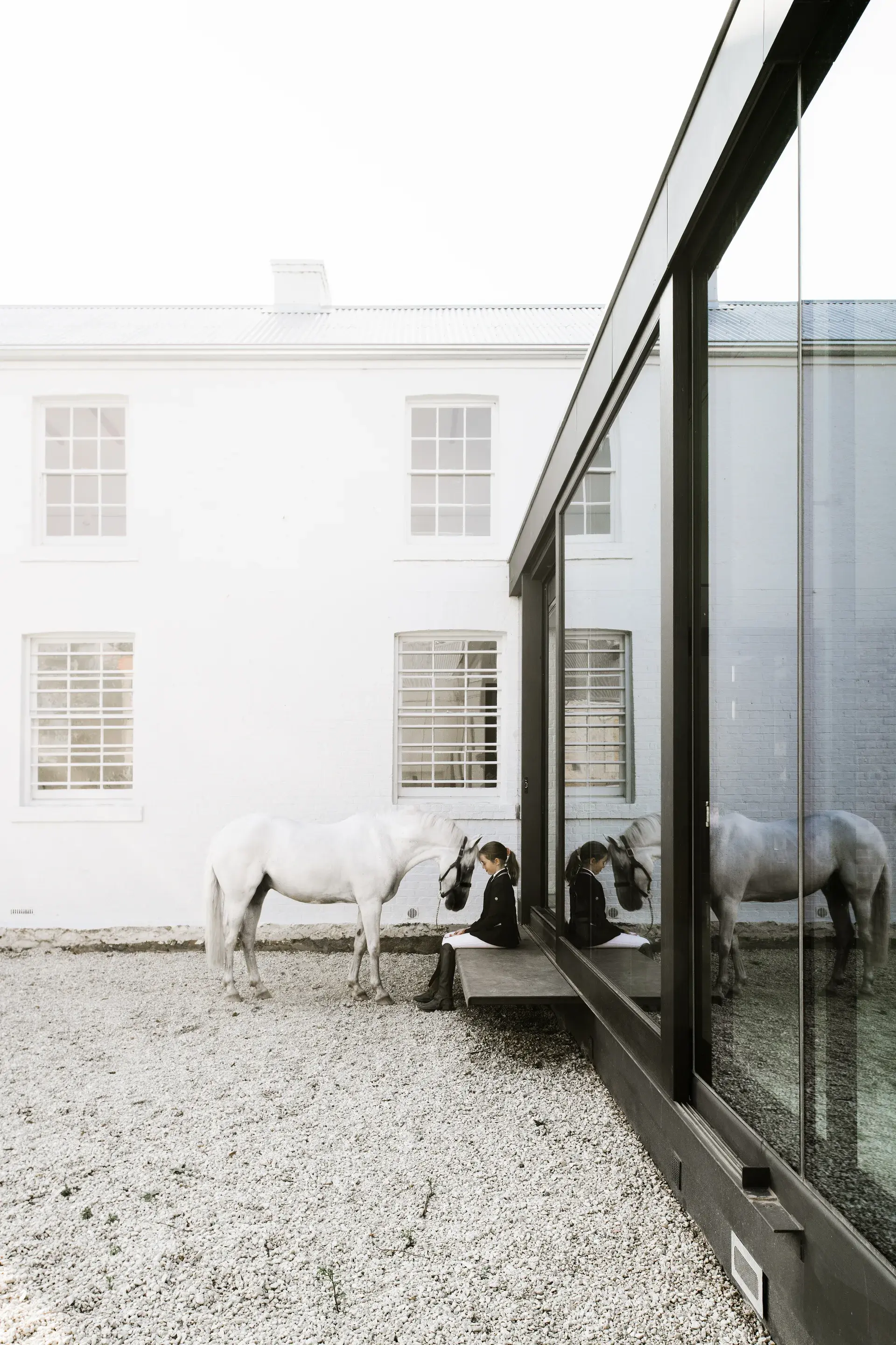 A young girl sits on a ledge beside a white horse, both reflected in the large glass windows of a modern building extension. Behind them stands a traditional two-story white building with a corrugated metal roof.