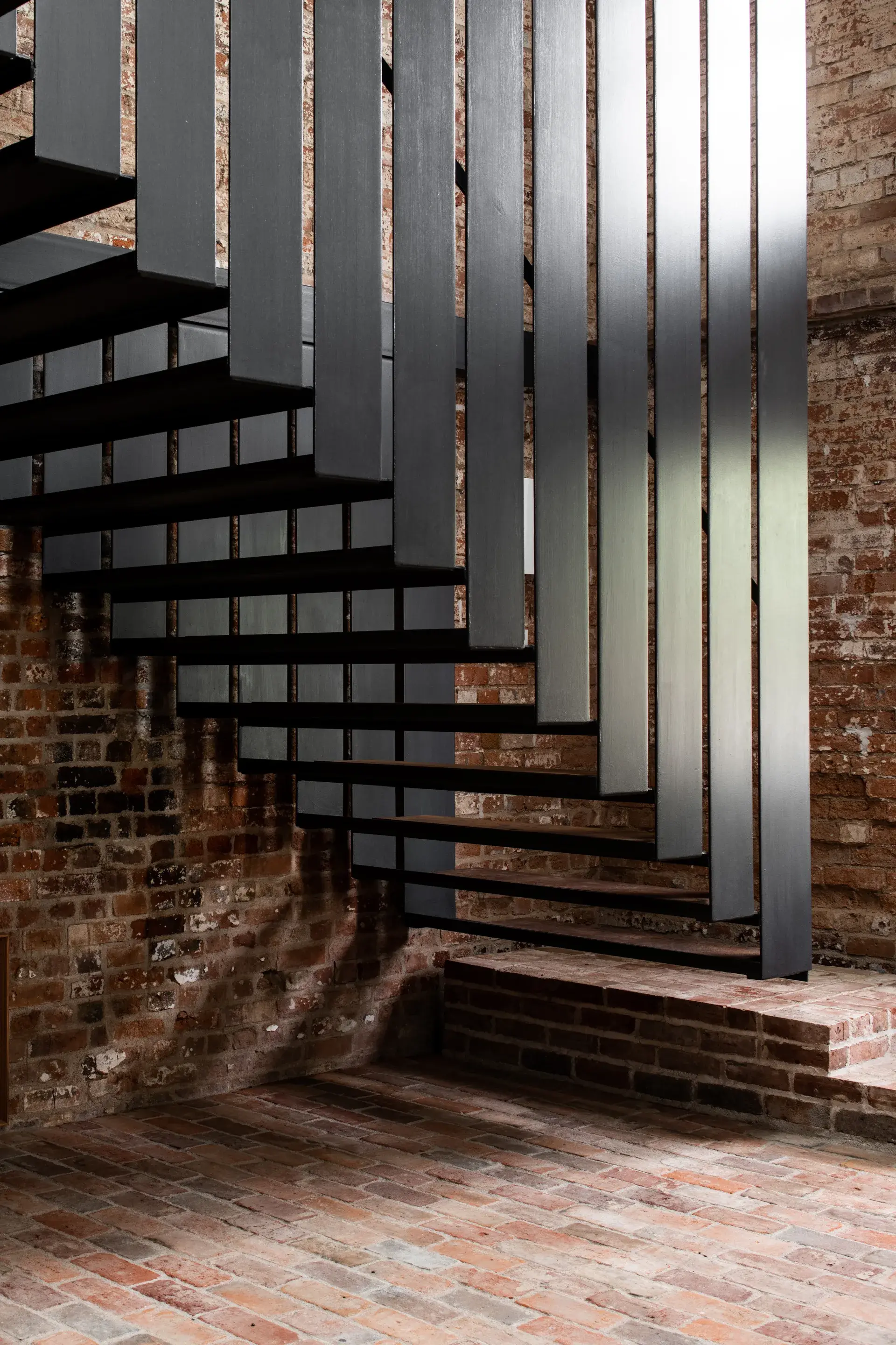 A staircase of dark metal treads and vertical slats appears to float against rustic exposed brick walls. The industrial metalwork creates a sharp contrast with the matching brick flooring.