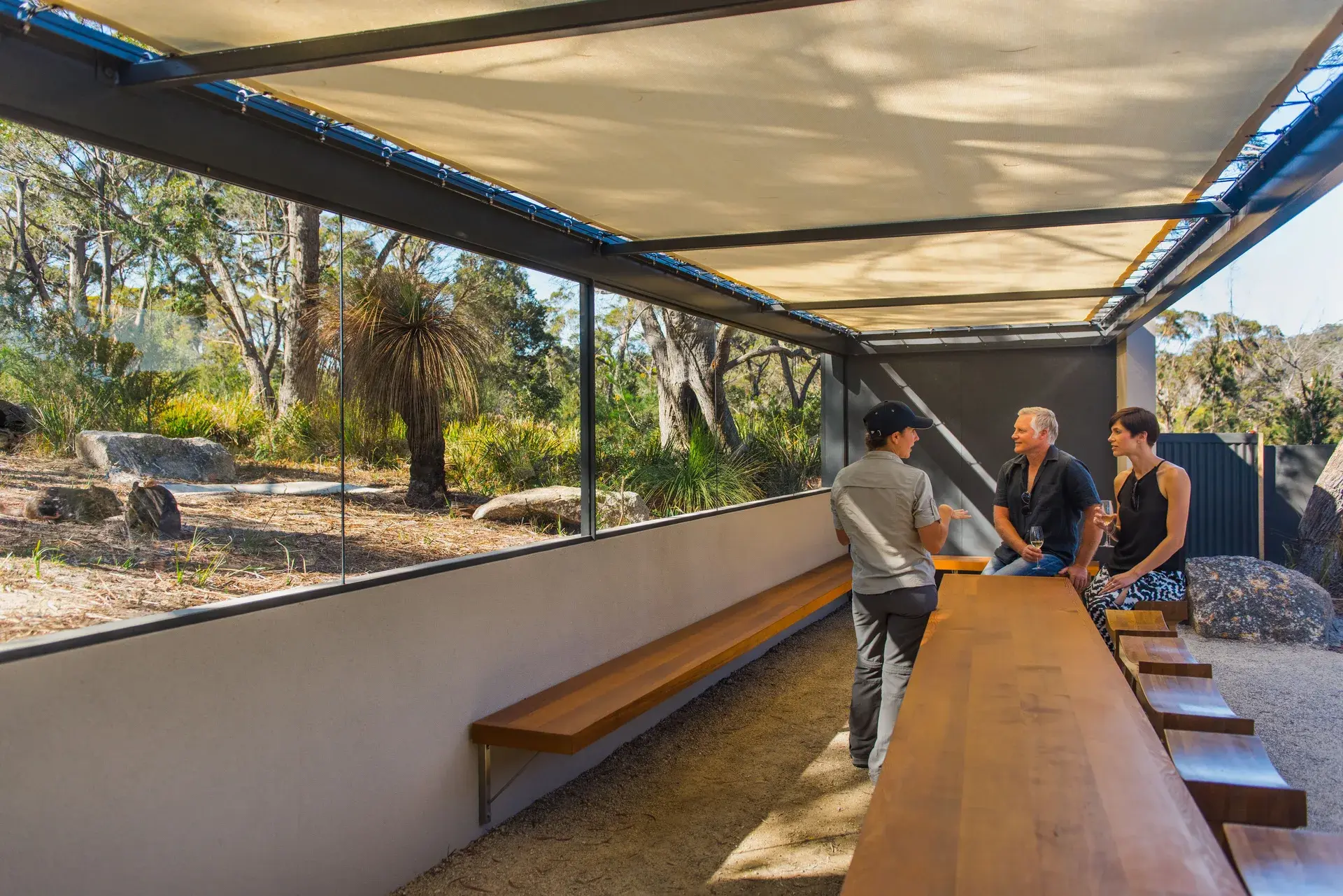 Three people sit at a long wooden table under a shaded canopy, engaged in conversation while holding glasses of wine. The seating area is enclosed by a large glass window that offers an expansive view of the surrounding forest and natural landscape.