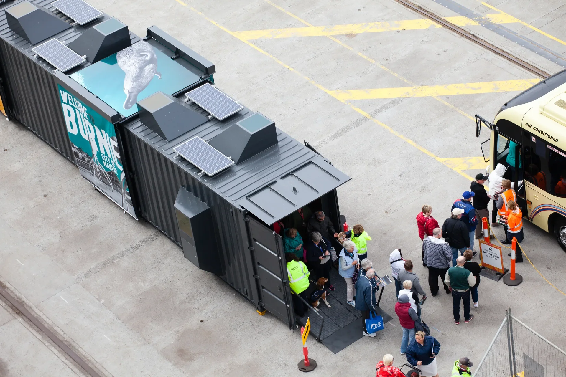 Cruise ship visitors in colourful clothing spilling out of the shelter and into the waiting tourist bus.