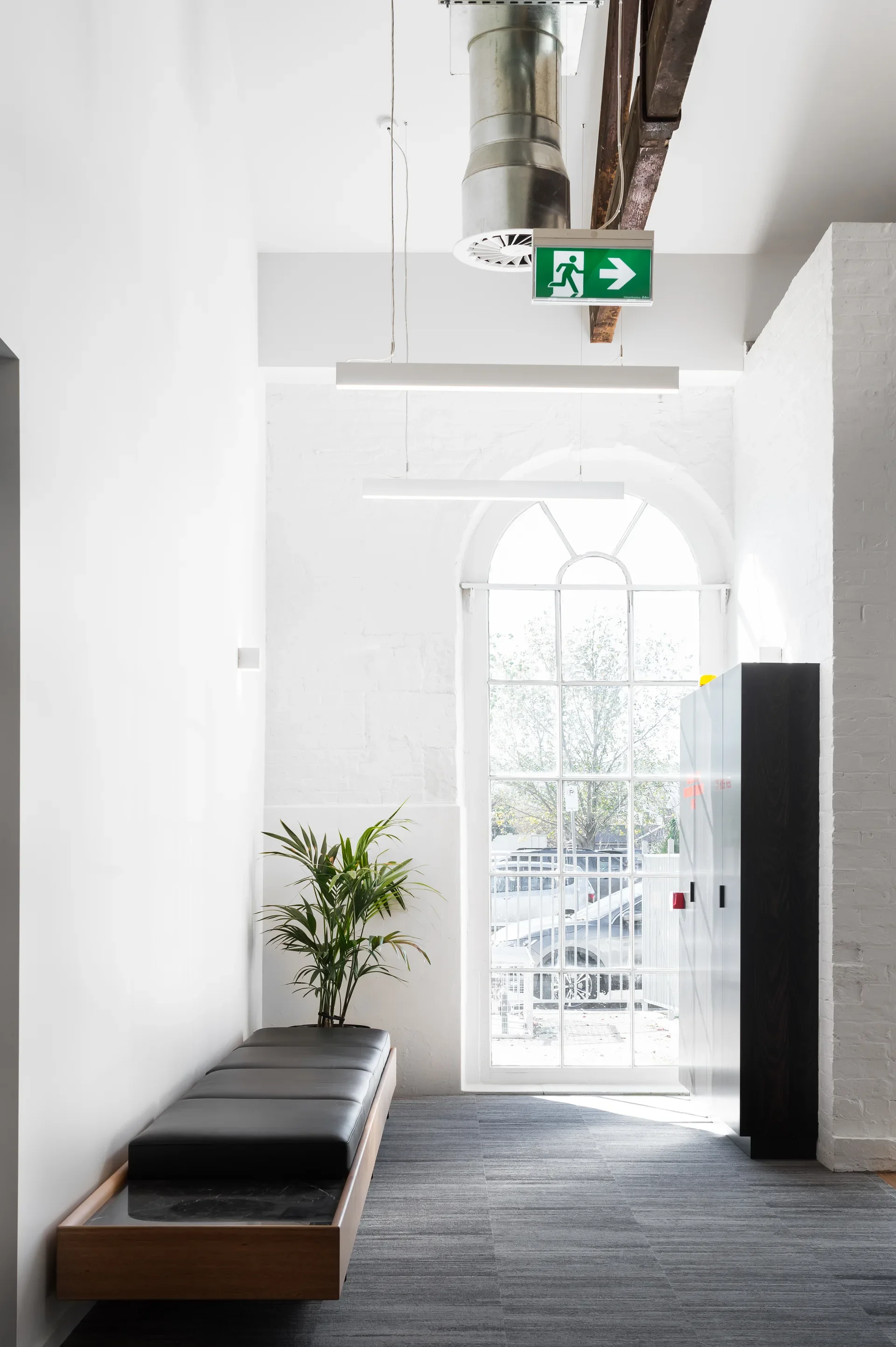 This bright waiting area features a black leather-cushioned bench with a wooden frame set against a clean white wall. A tall arched window provides a view of the outdoors, while the industrial ceiling includes exposed wooden beams and a metal ventilation duct.