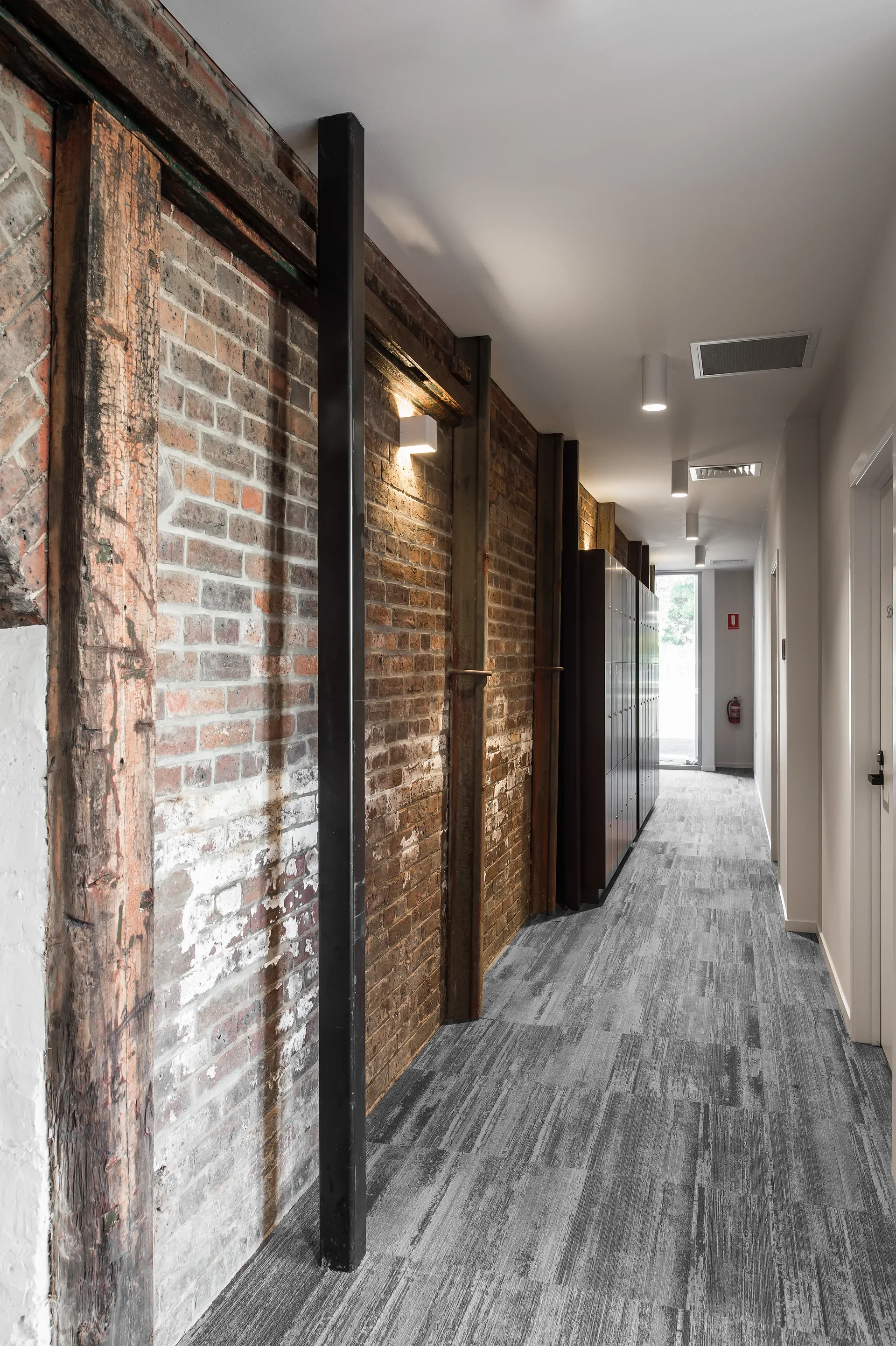 This long office corridor showcases a textured wall of original exposed brick and heavy timber beams illuminated by spotlights. A row of dark lockers and grey patterned carpet tiles lead the eye toward a bright window at the far end.