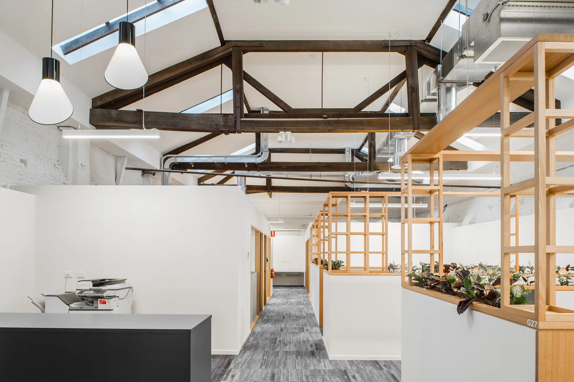 This open-plan office features vaulted ceilings with dark wood trusses and integrated skylights. White partition walls are topped with light wood lattice shelving and greenery, leading past a black service station toward a long corridor.