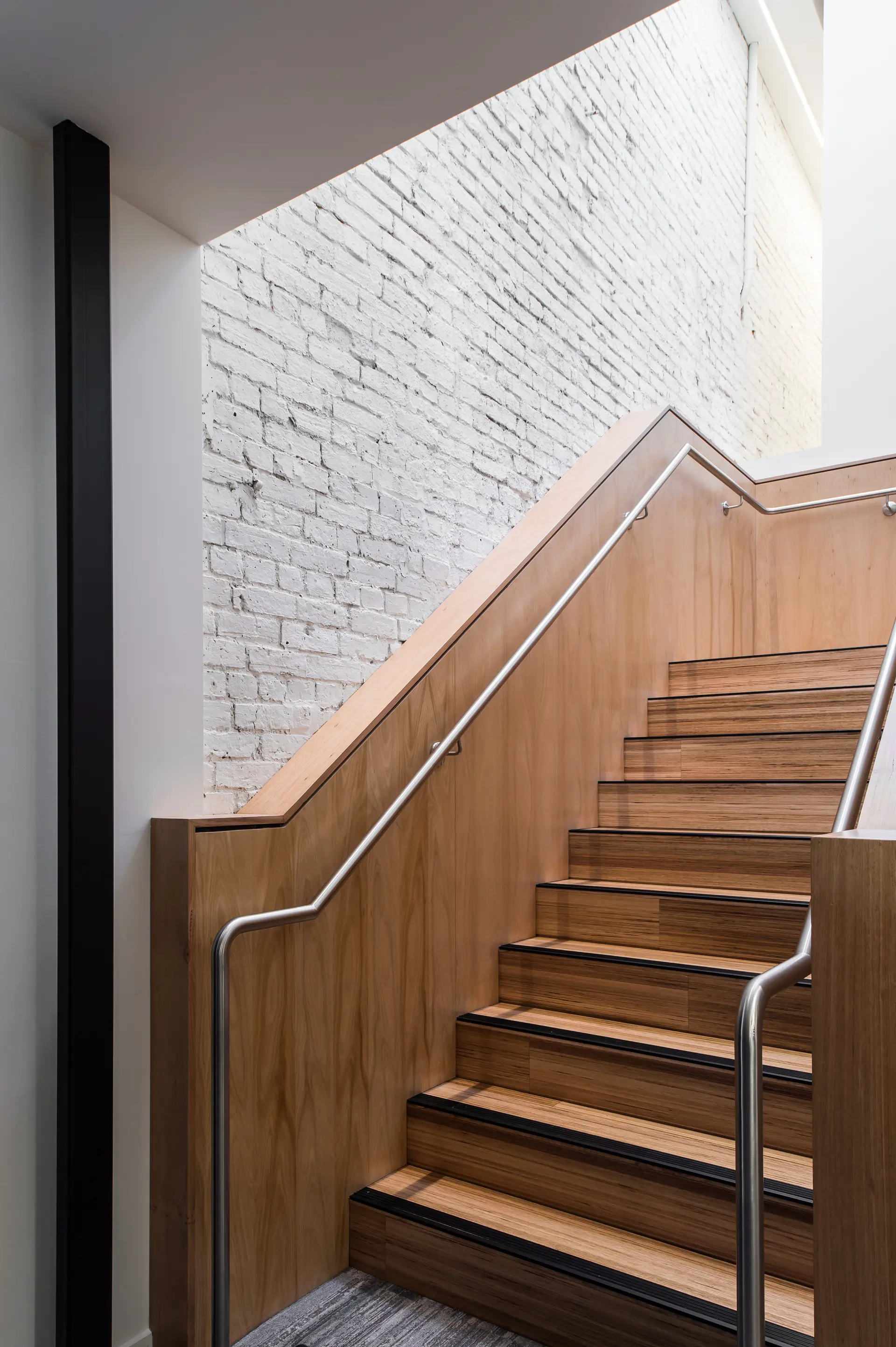 This modern staircase features natural wood-panelled walls and steps with non-slip black edges. It is paired with a sleek, continuous metal handrail and set against a white-washed brick wall that extends toward a bright upper level.