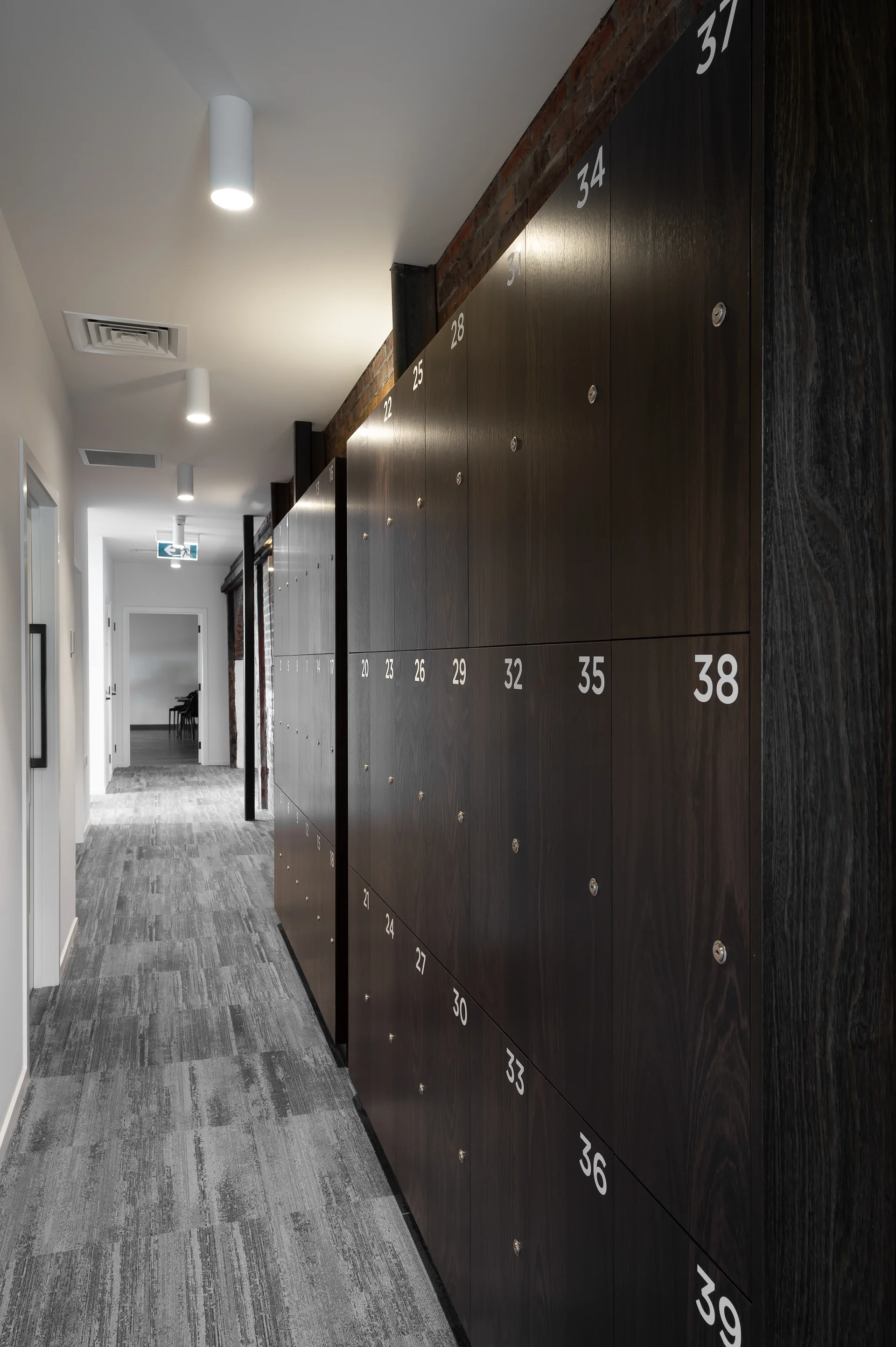 This office corridor features a long bank of numbered dark wood lockers stretching along the right-hand wall. The hallway is finished with grey patterned carpet and cylindrical white ceiling lights, leading toward an open doorway at the far end.