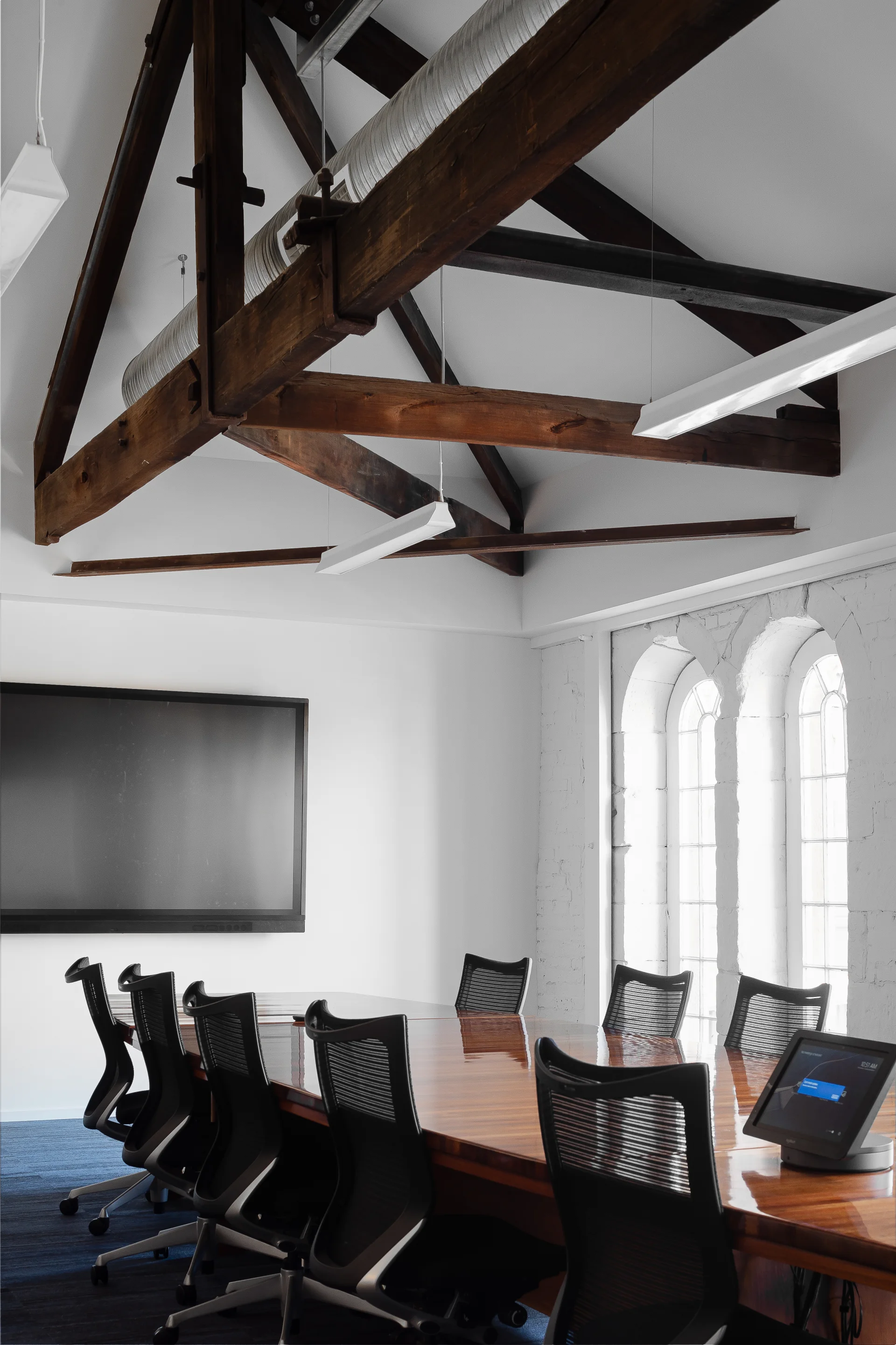 This boardroom features a polished wooden table with black mesh chairs under massive dark wood ceiling trusses. The space is equipped with a large wall monitor and a tabletop touchscreen, set against a white brick wall with arched windows.