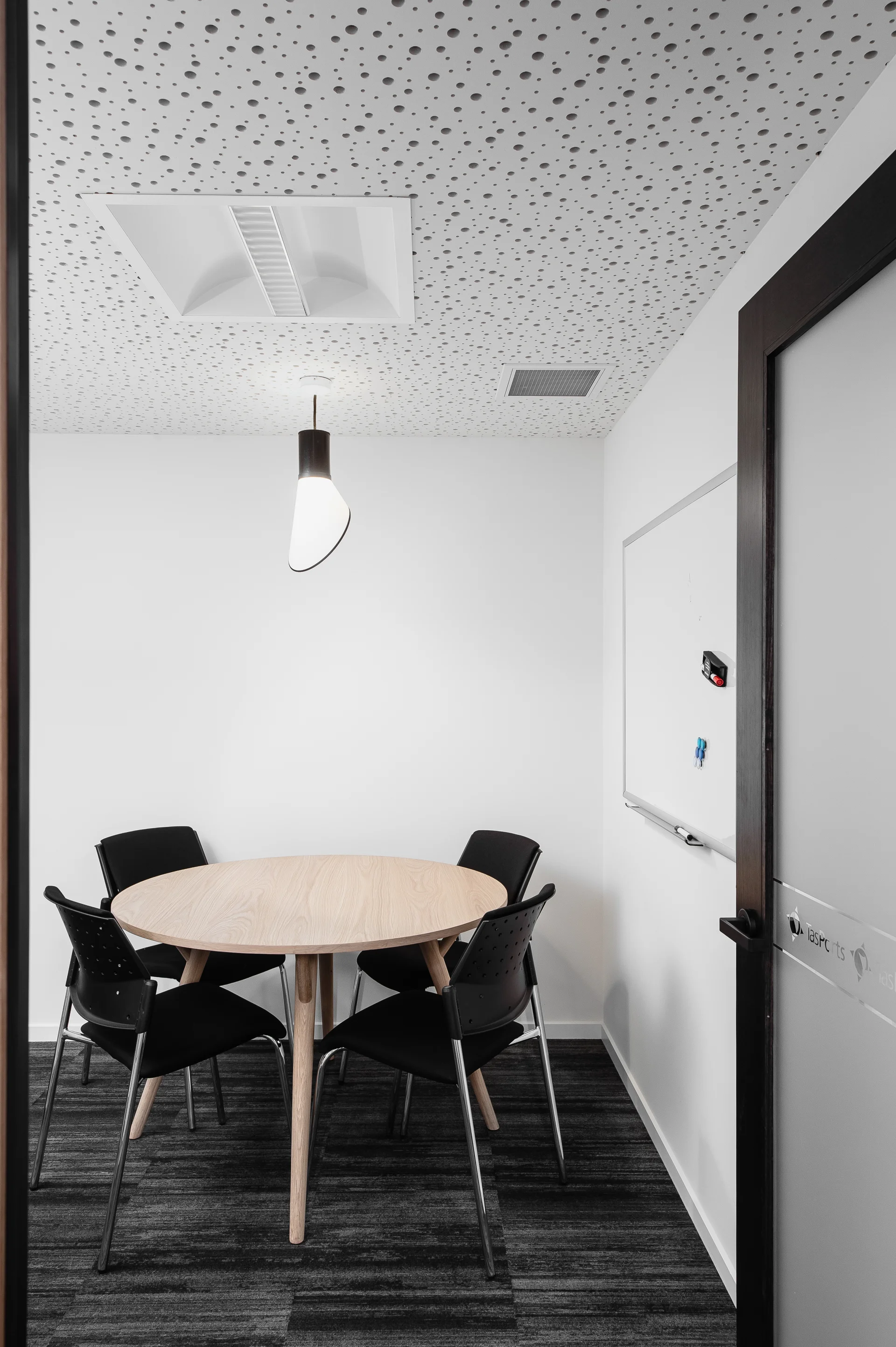 Gemini said This small meeting room features a light wood circular table surrounded by four black chairs on a grey patterned carpet. The clean white walls are accented by a whiteboard and a unique, curved pendant light hanging from a distinctive perforated acoustic ceiling.