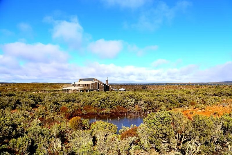 The large timber lodge is situated in a rugged, low-shrub landscape under a bright blue sky with scattered clouds. A small pond sits in the foreground, reflecting the surrounding dense greenery and the modern, slanted roofline of the building.