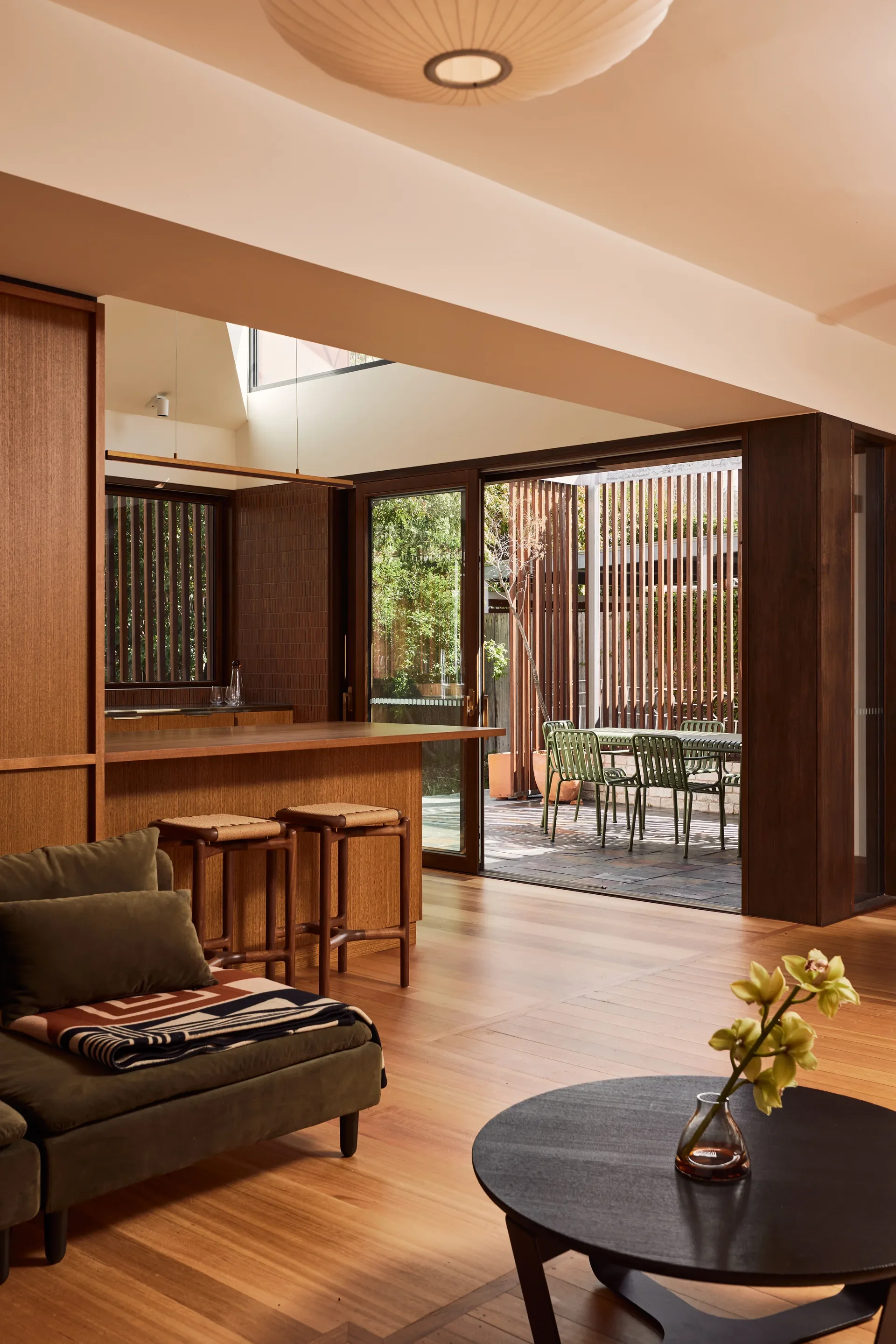 This warm, modern living area features rich timber paneling and polished wood floors that extend toward an outdoor patio. Sliding glass doors reveal an outdoor dining set framed by vertical timber slats, while the interior is furnished with a dark green lounge and a circular black coffee table.