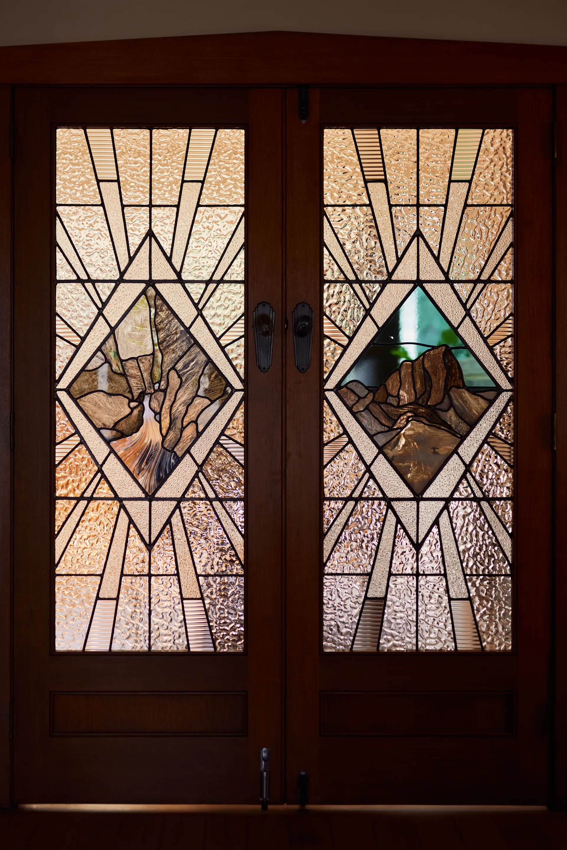 These wooden double doors feature intricate leadlight glass panels with an Art Deco geometric design. Central diamond segments depict stylized mountain landscapes surrounded by textured amber and clear glass sunburst patterns.