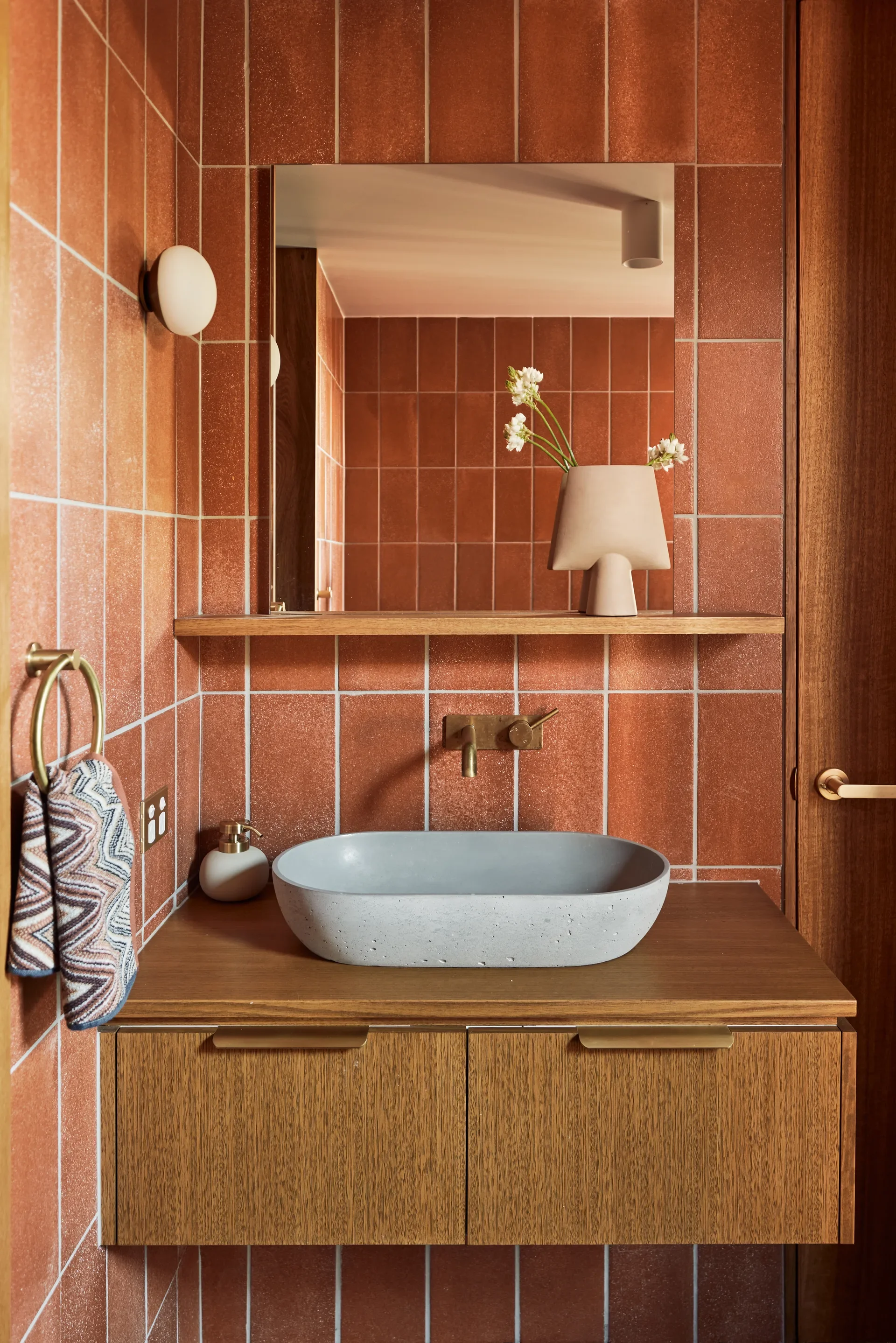 This modern bathroom features terracotta-coloured vertical tiles and a floating wooden vanity. It is equipped with a grey concrete vessel sink, brass hardware, and a large mirror that reflects a unique beige vase with white flowers.