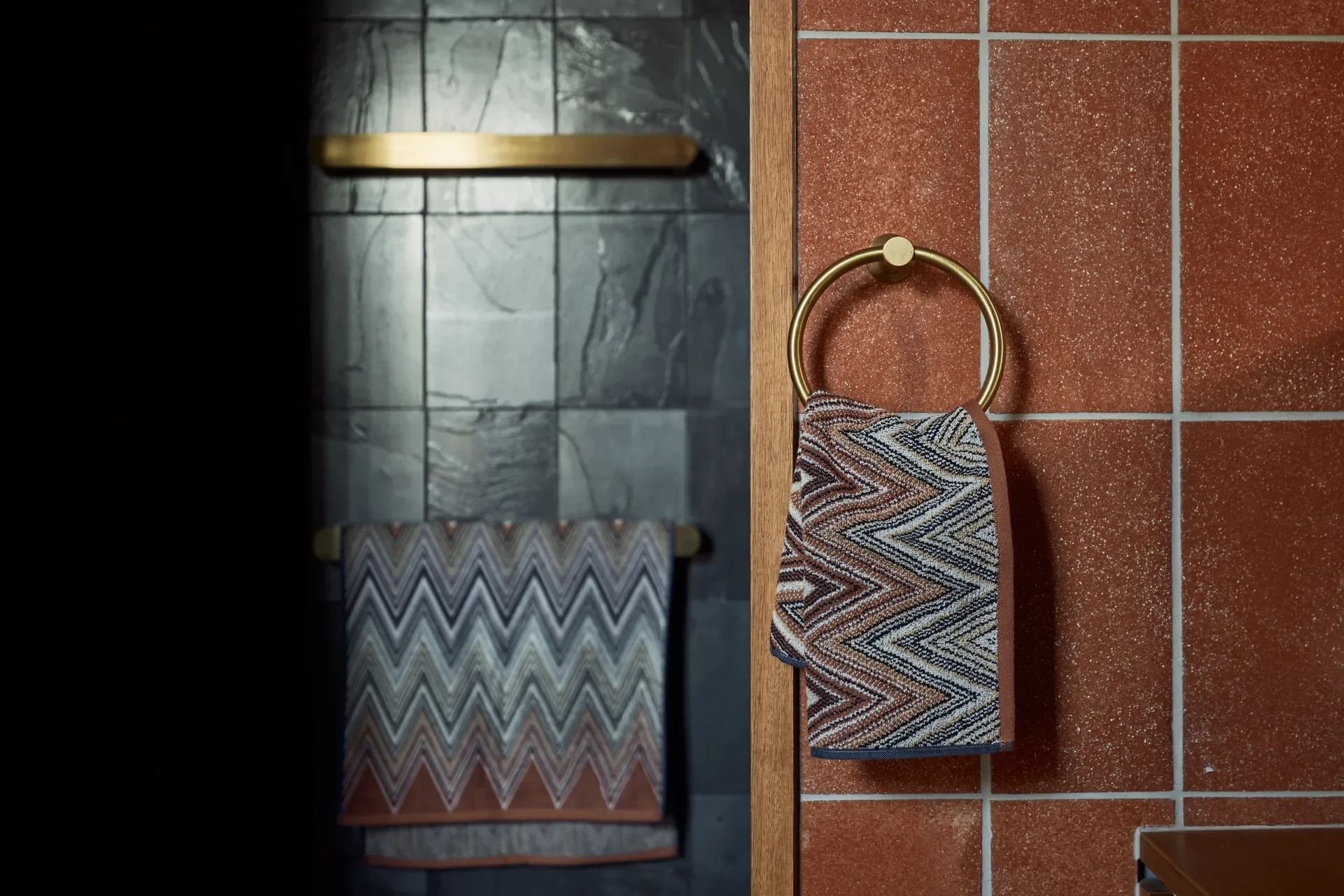 This bathroom detail features terracotta-toned tiles paired with brass hardware, including a circular towel ring. A patterned zigzag towel hangs in the foreground, while the background reveals a dark, slate-tiled shower area accented by a horizontal brass shelf.