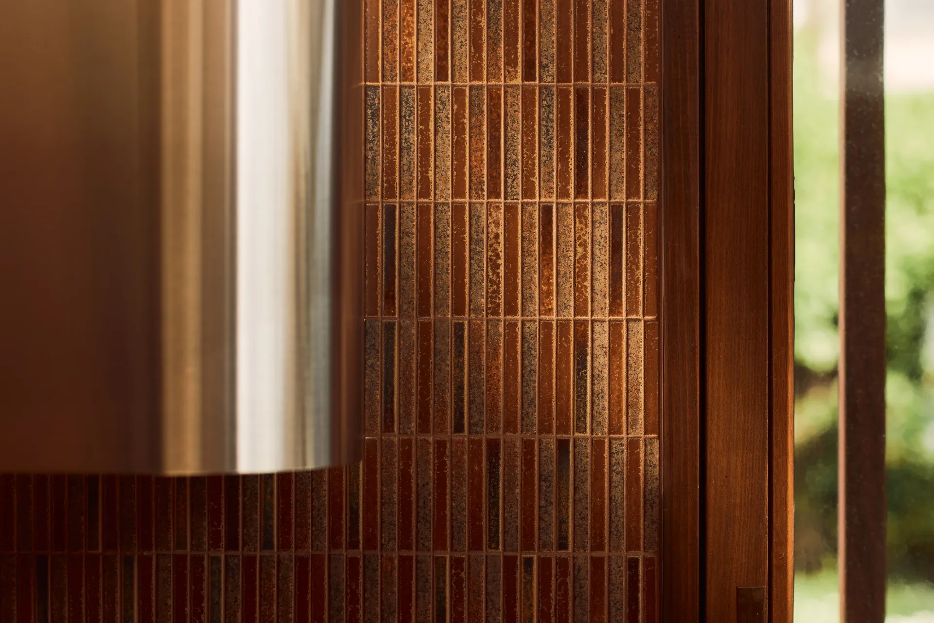 This kitchen detail features a wall of narrow, vertical tiles in a rich, textured brown-orange hue. The tiling is positioned next to a dark wood window frame and is partially obscured by the curved, metallic surface of a stainless steel range hood.