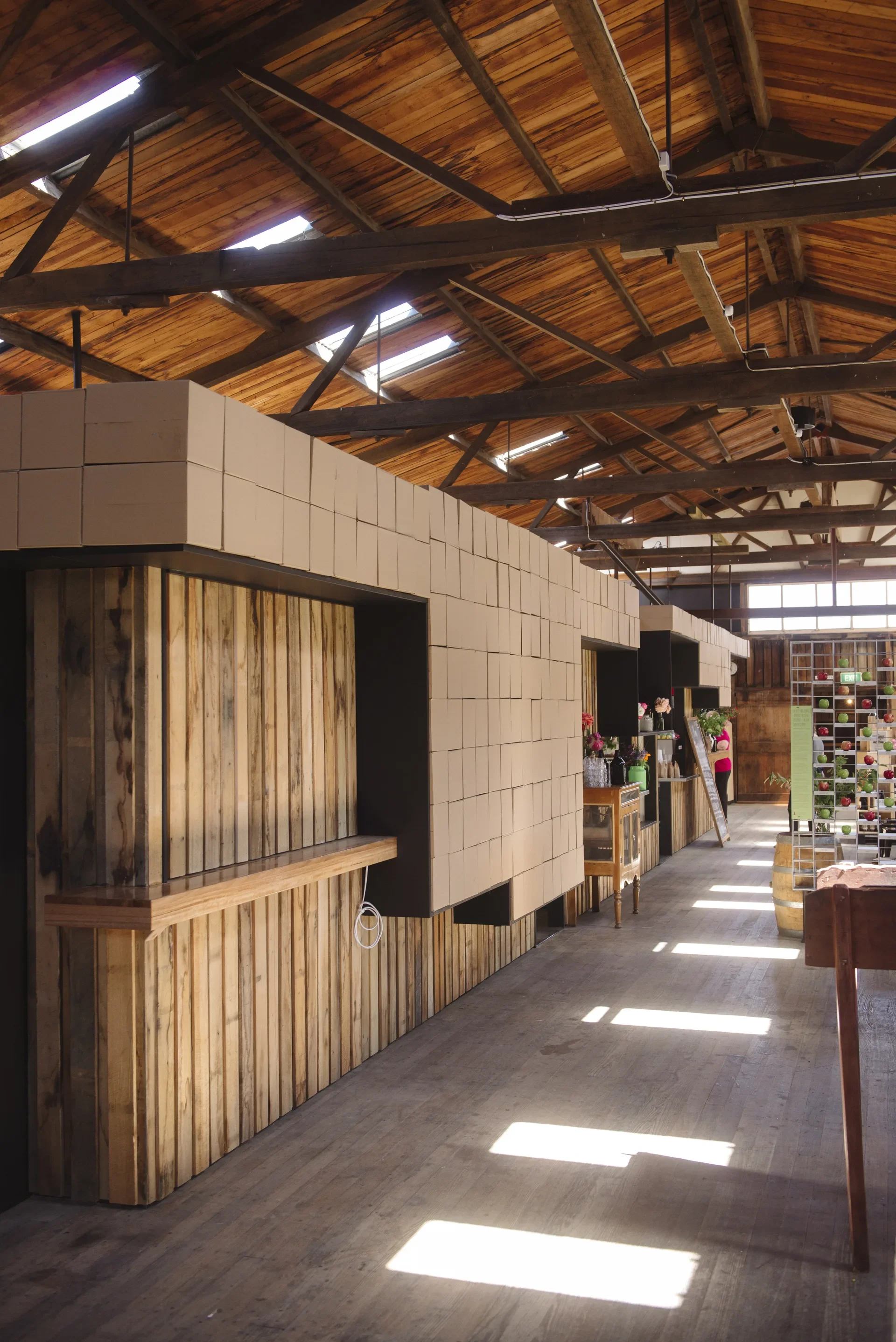 This rustic interior features a vaulted timber ceiling with exposed beams and small skylights. The space is divided by long counters clad in vertical wood planks, topped with an arrangement of cardboard boxes..
