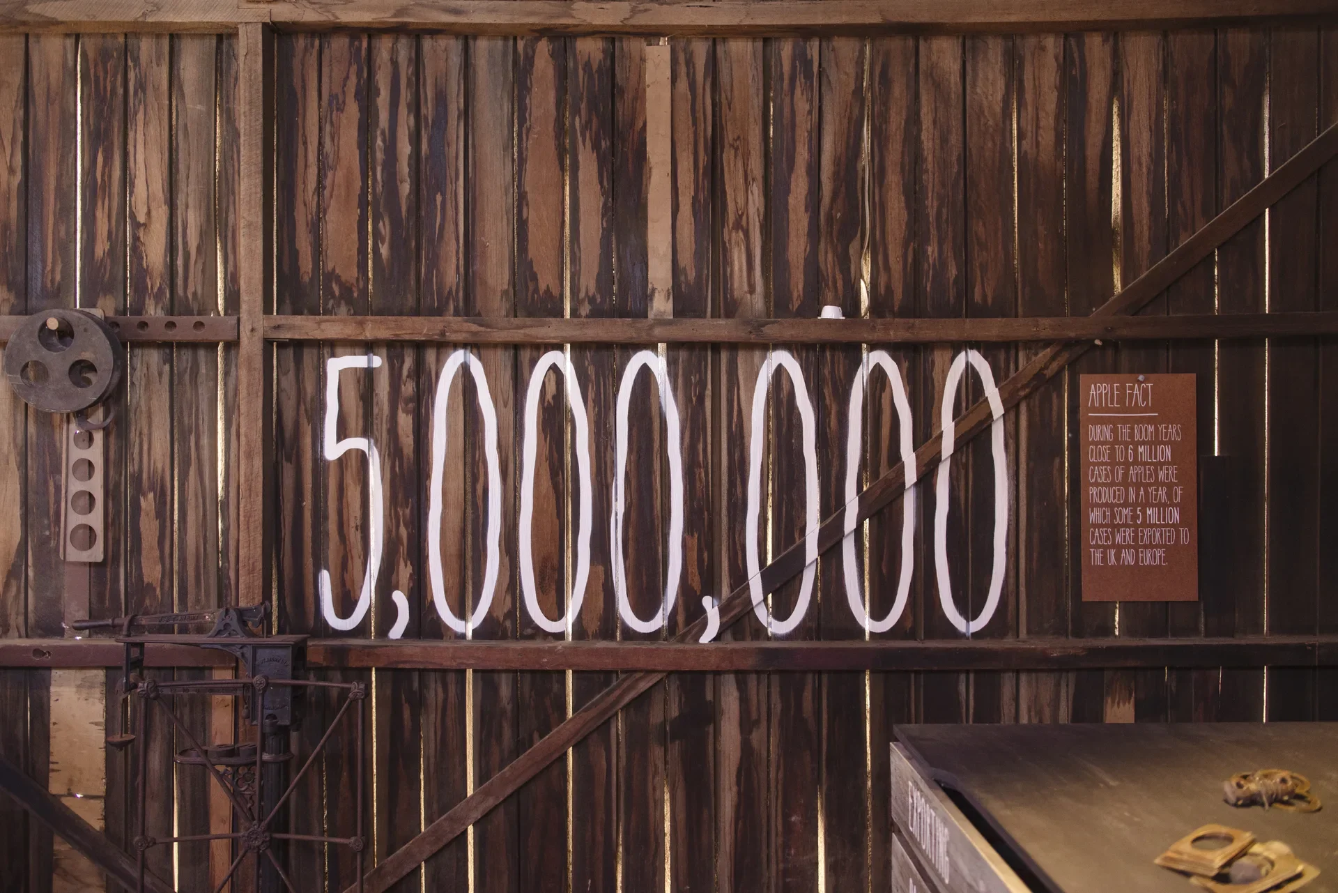 This rustic museum display features dark, weathered vertical timber planks with a large hand-painted "5,000,000" in white. Beside the number, an "Apple Fact" plaque explains that during boom years, 5 million cases of apples were exported to the UK and Europe.