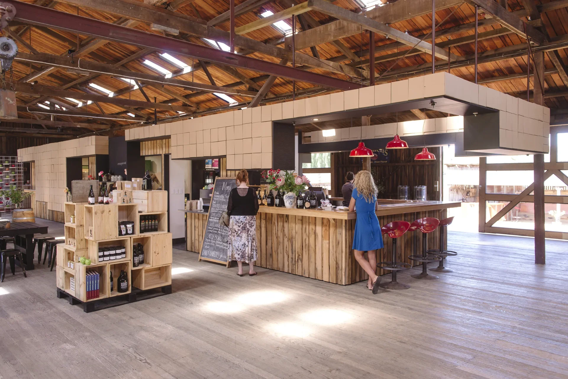 This rustic interior features a vaulted timber ceiling with exposed beams and skylights. A long service counter clad in vertical wood planks is topped with bright red pendant lights, while nearby products are displayed on a tiered unit made from stacked wooden crates.