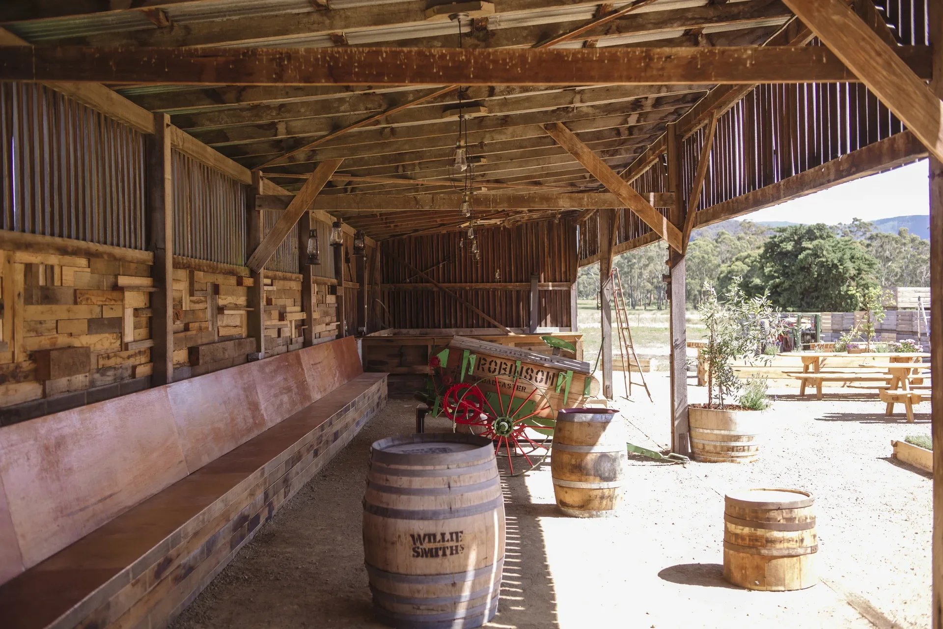 This open-sided timber shed features a long wooden bench and rustic cider-making artifacts. Several branded "Willie Smiths" barrels and a vintage "Robinson" machine with red wheels are positioned under the exposed-beam roof.