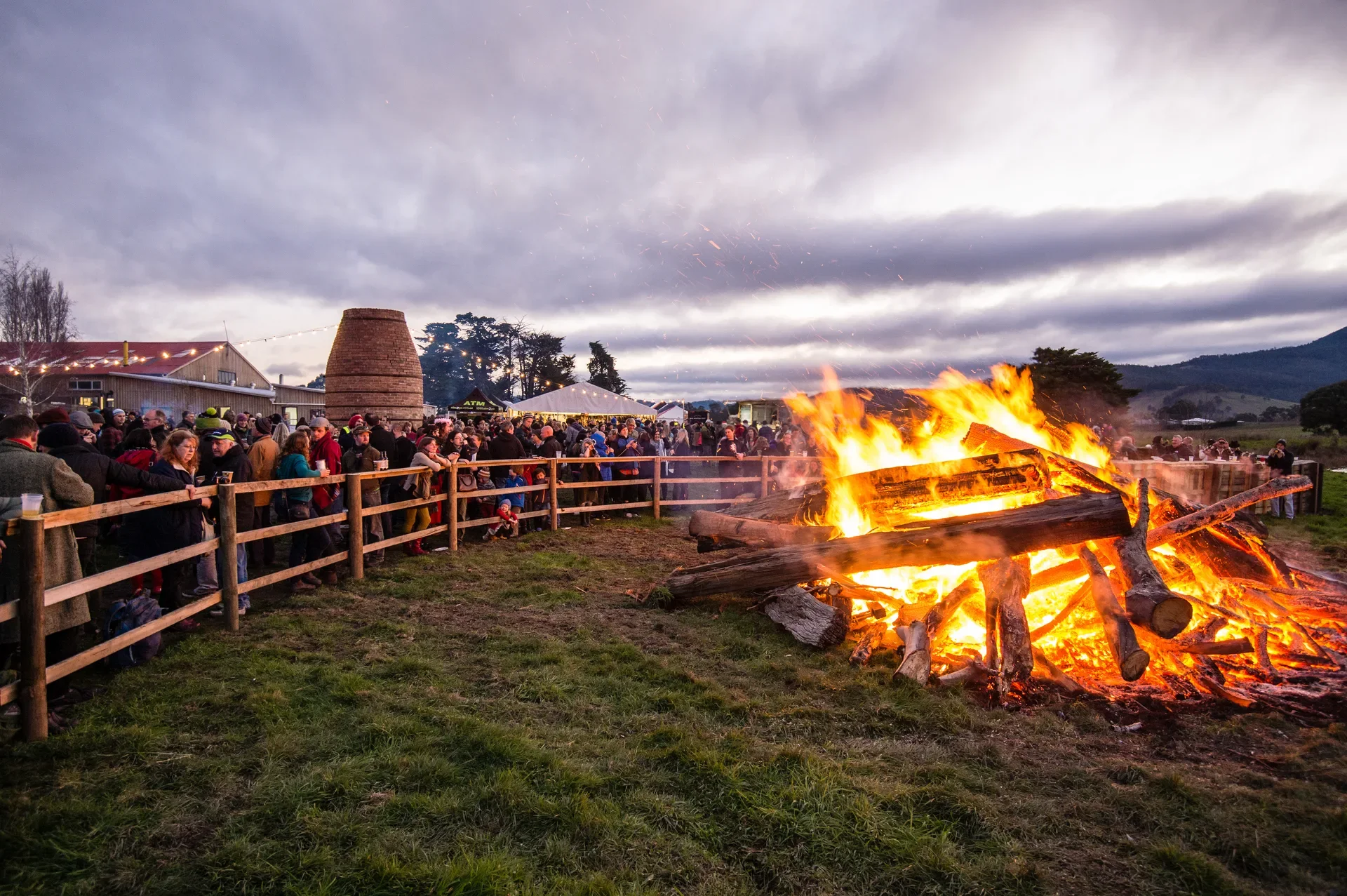 A large, roaring bonfire of thick logs casts a bright orange glow over a crowd gathered behind a wooden fence. A tall brick kiln-like structure and event tents are visible in the background under a cloudy twilight sky.