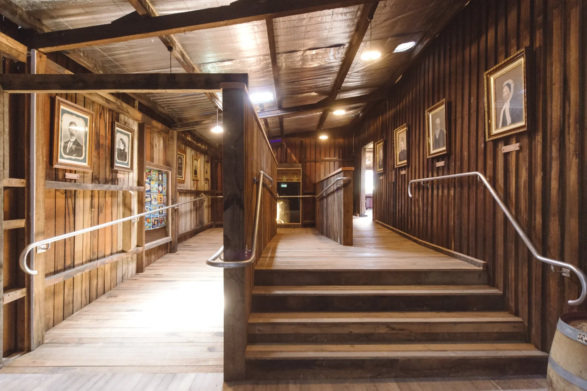This rustic gallery space features a wooden staircase and a ramp with silver handrails, leading into a hallway lined with framed historical portraits. The interior is constructed with vertical timber planks and has a low-slung ceiling with exposed beams and simple overhead lighting.