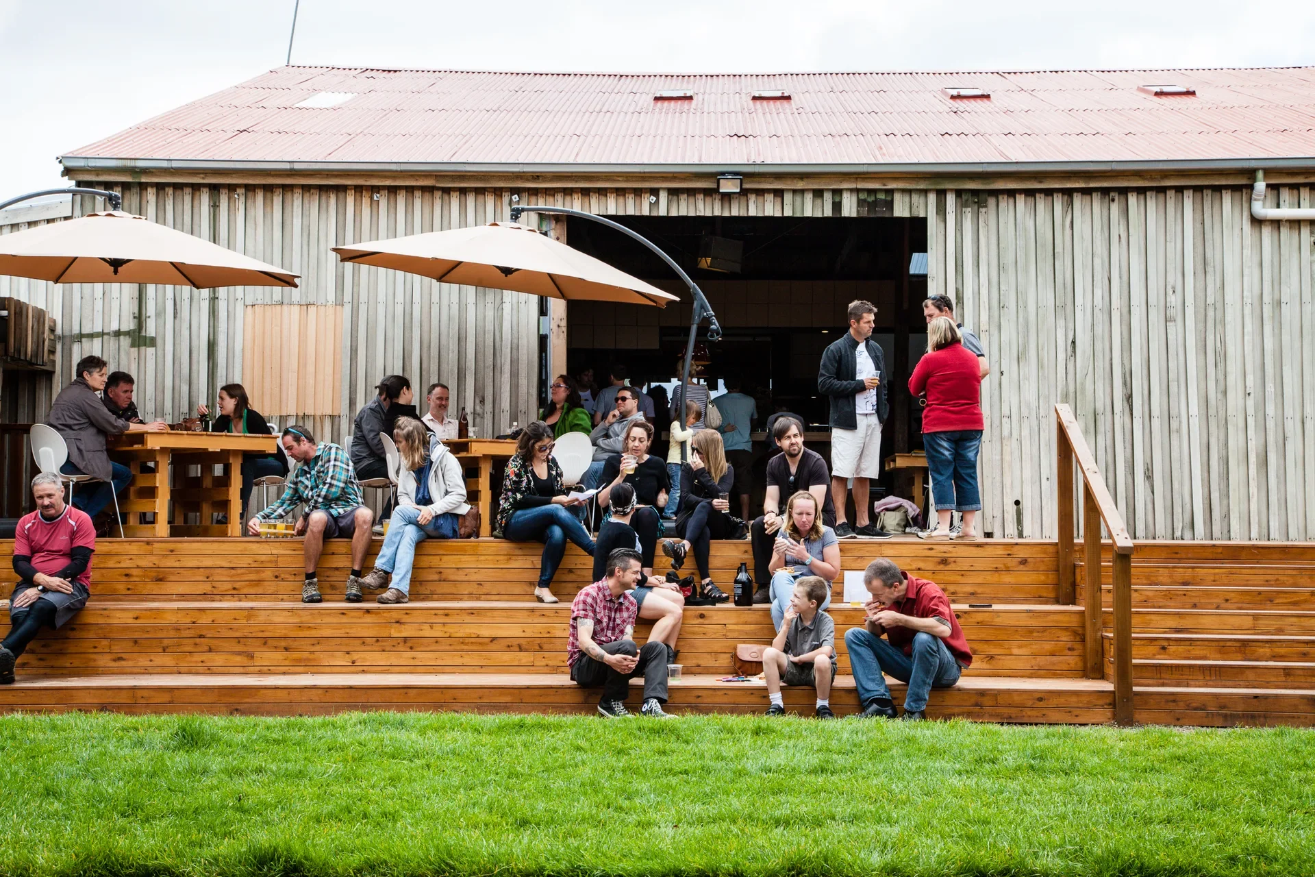 A crowd of people relaxes on a wide, multi-tiered wooden deck in front of a large timber building with a red roof. Some visitors sit at tables under large tan umbrellas, while others lounge on the steps overlooking a bright green lawn.