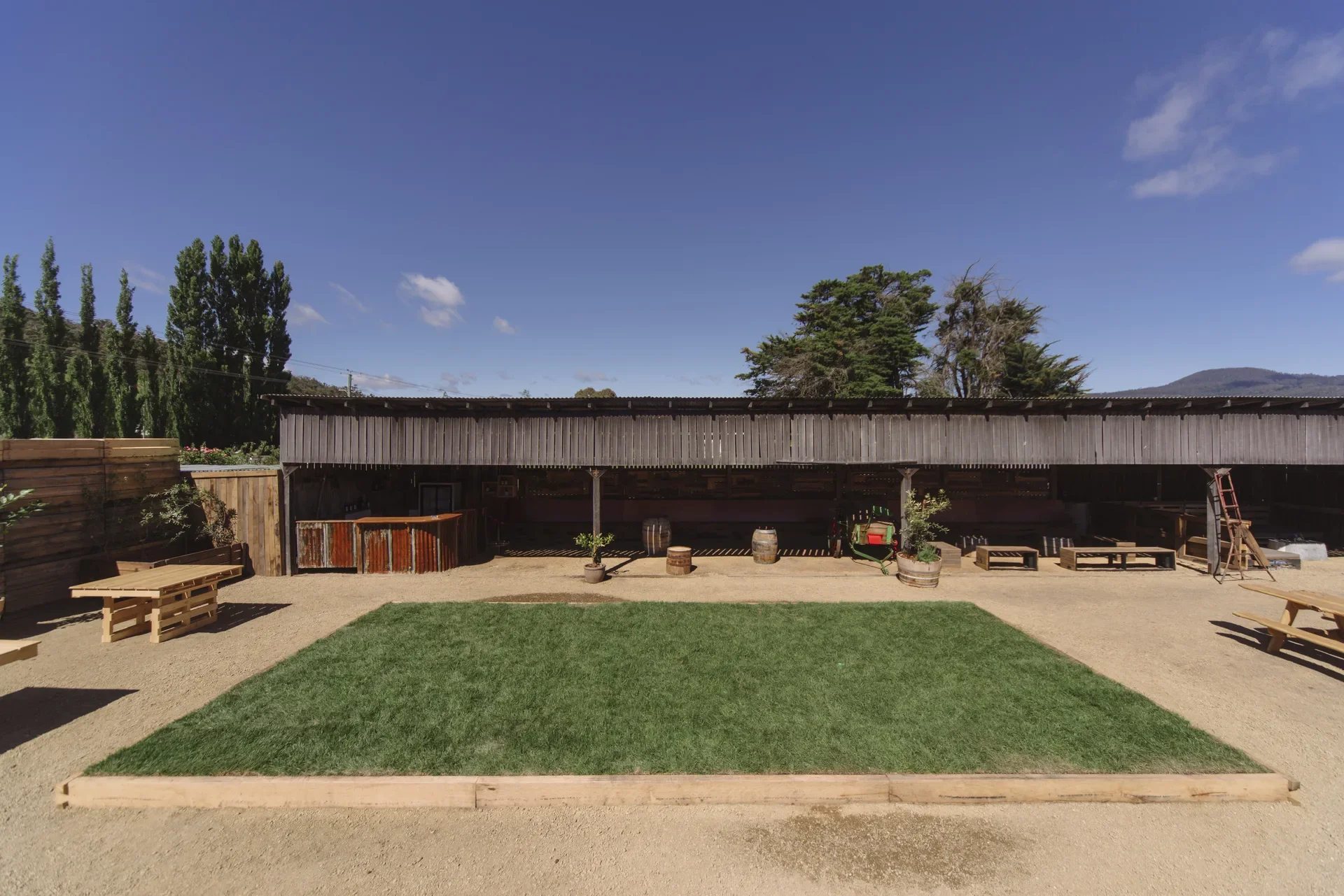 This outdoor space features a rectangular patch of green lawn centered in a wide, gravel courtyard. In the background, a long, low-slung timber shed provides a shaded area with a corrugated metal bar and scattered wooden seating.
